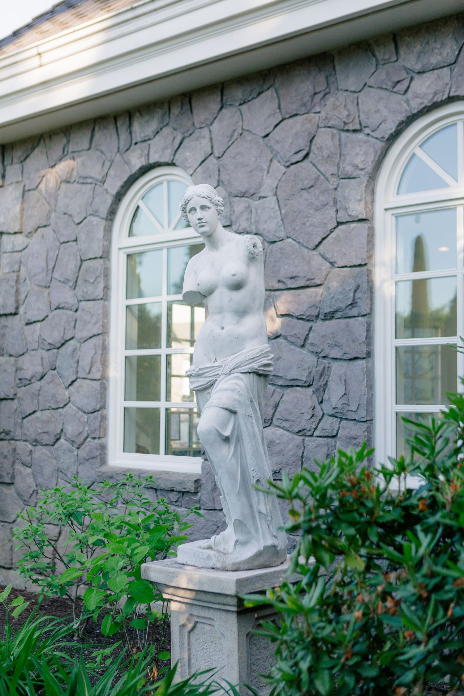 Classical Venus-style garden statue stands between arched windows on a stone wall with lush greenery around the base.