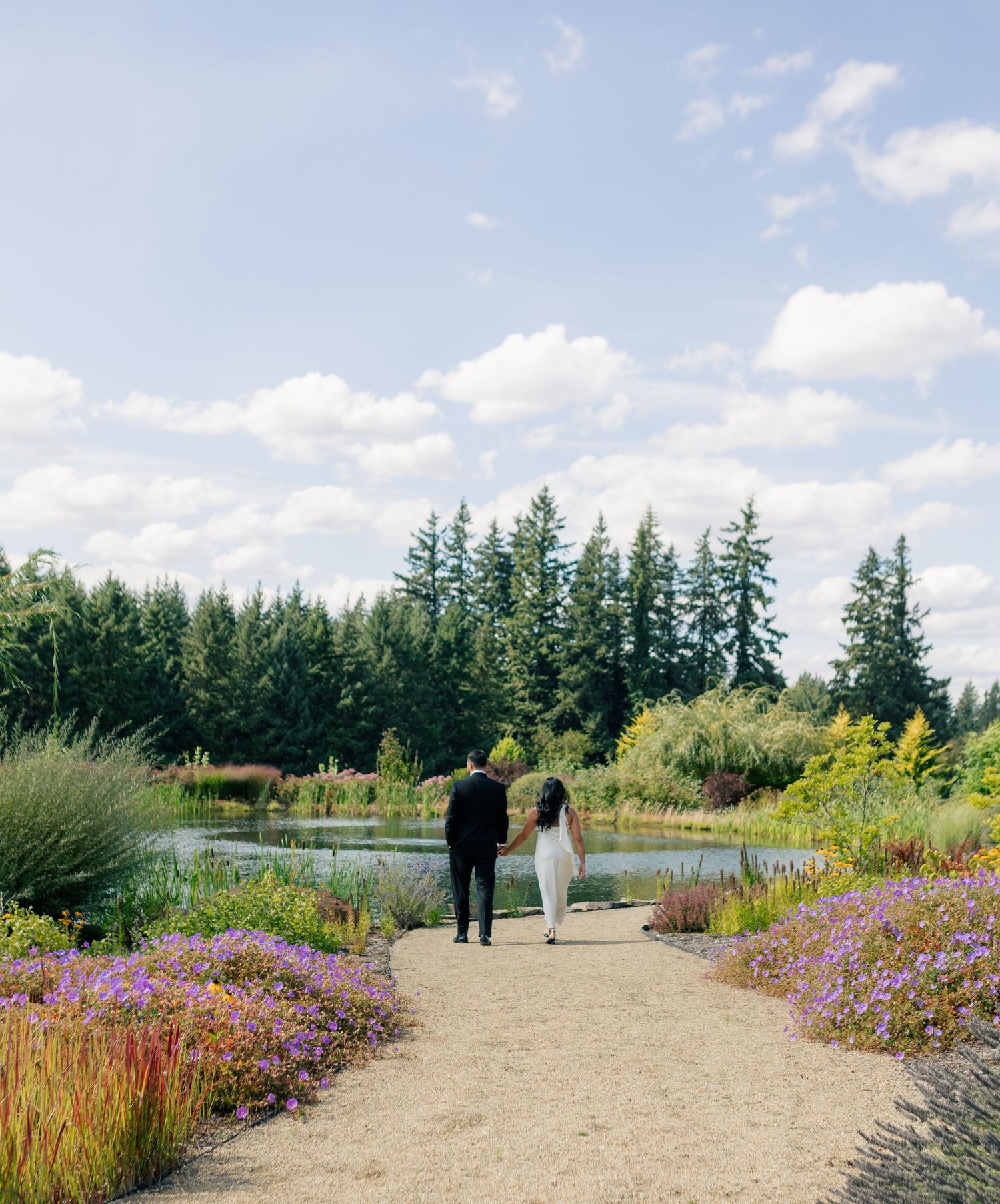 Gravel path winding through colorful garden beds with purple flowers and evergreen trees under blue sky.