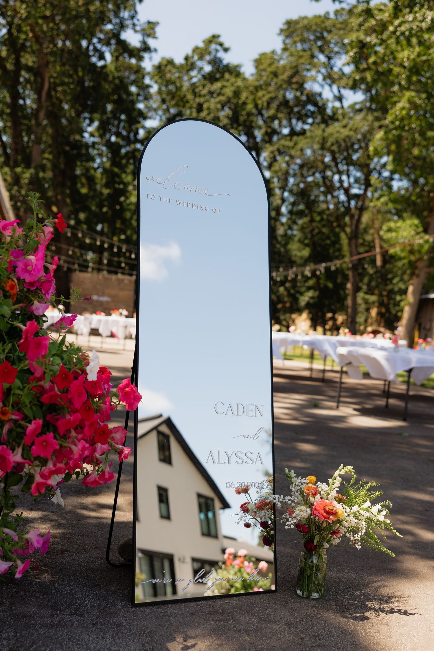 Elegant arched mirror wedding welcome sign for Caden and Alyssa outdoors, surrounded by colorful floral arrangements.