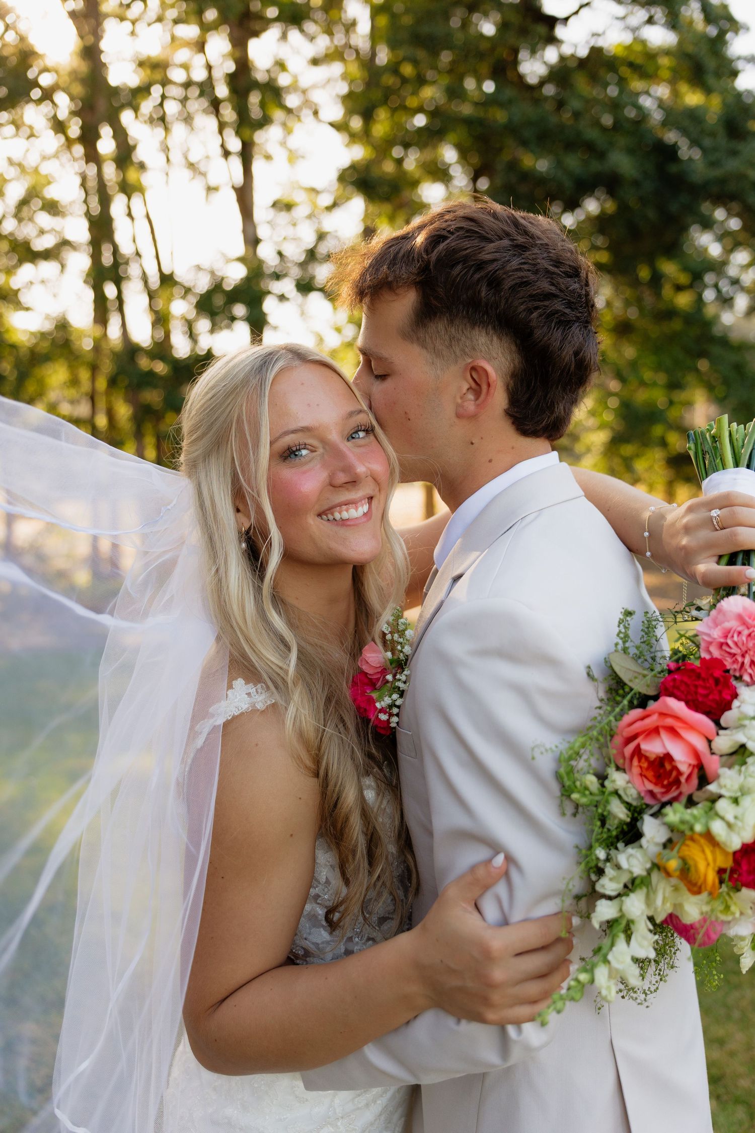 Smiling bride with flowing veil held by groom in cream suit holding colorful bridal bouquet.