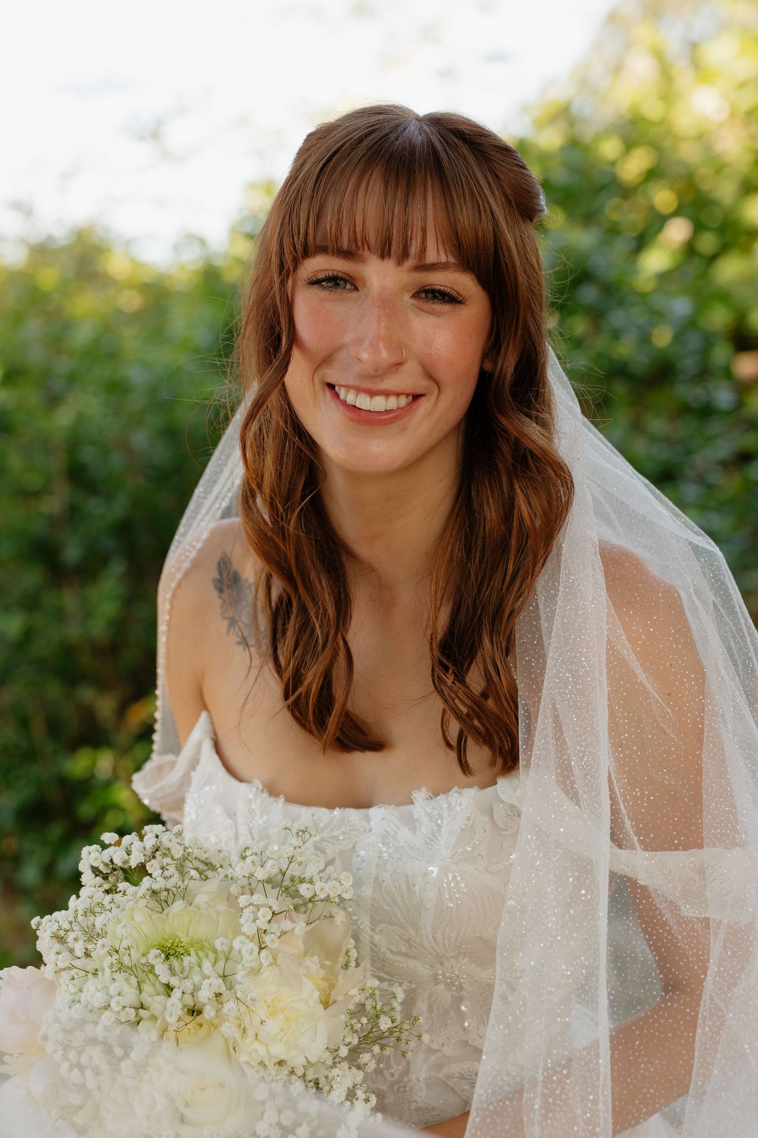 Smiling bride with bangs wearing a veil and holding a white floral bouquet in a sunny outdoor setting.