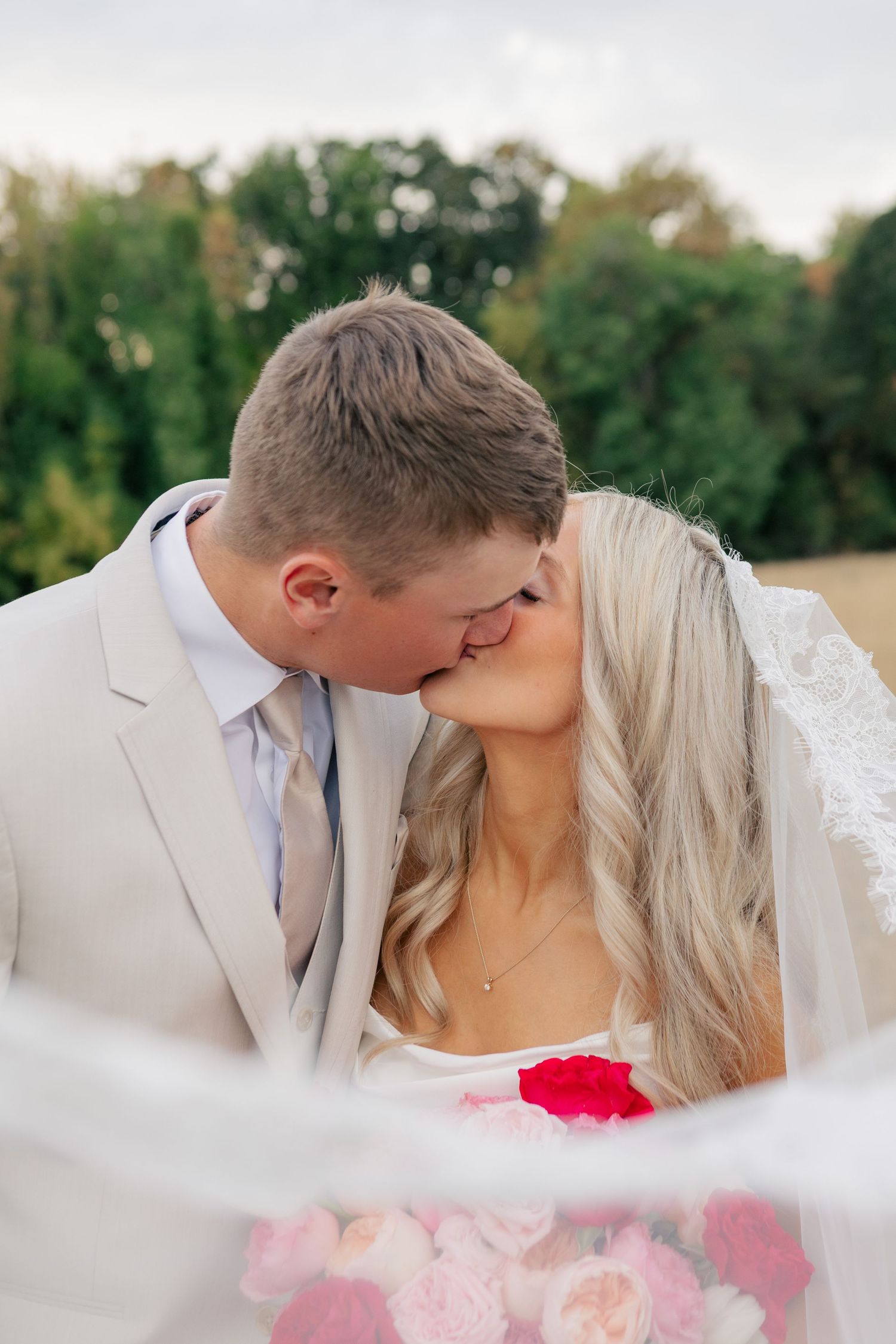 Bride and groom sharing a romantic kiss outdoors on their wedding day, with a floral bouquet and veil in the foreground.
