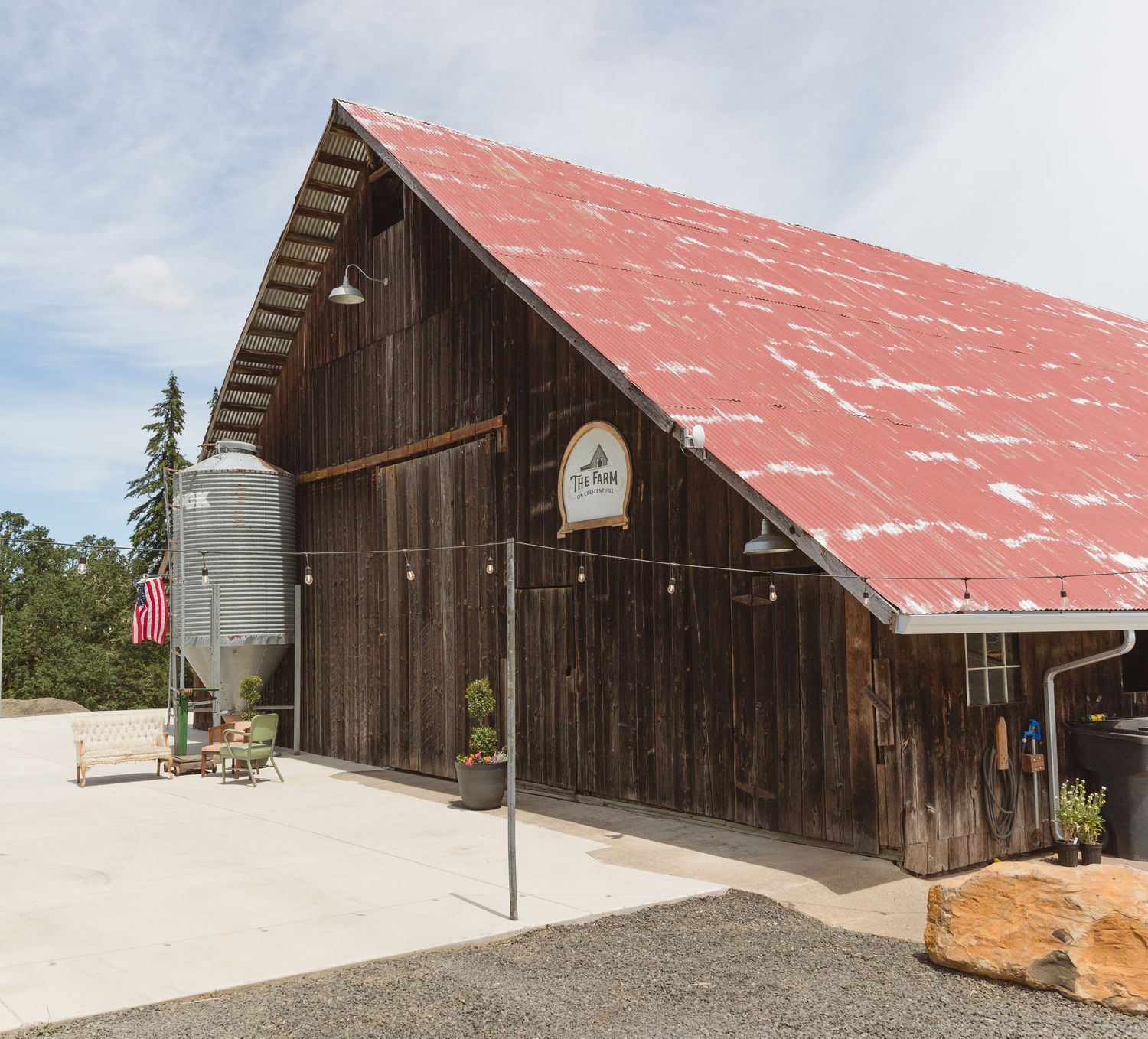 A large rustic barn with weathered wood siding, red metal roof and grain silo against a cloudy sky.