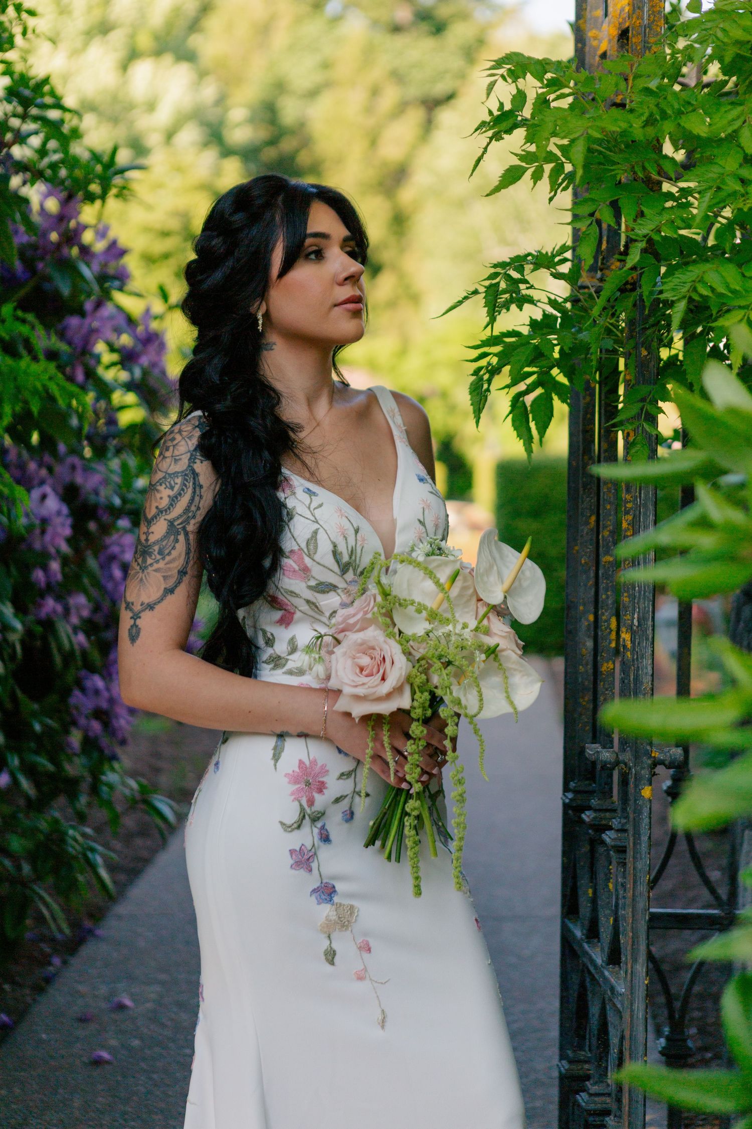 A bride in a floral embellished white wedding dress holds a calla lily bouquet while standing near purple flowers in a garden.