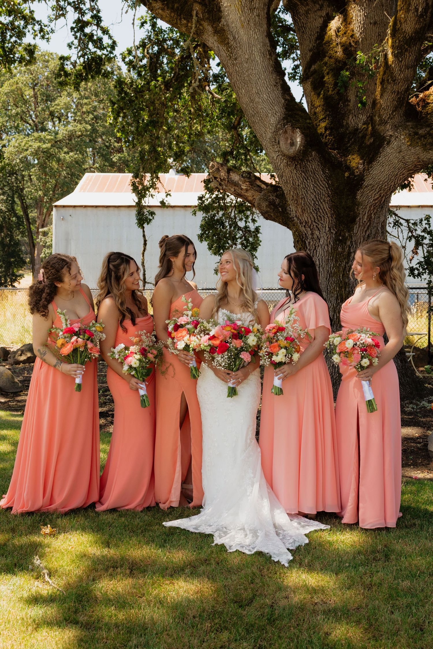 Bride in lace gown poses with five bridesmaids in coral dresses holding bouquets under large oak tree.