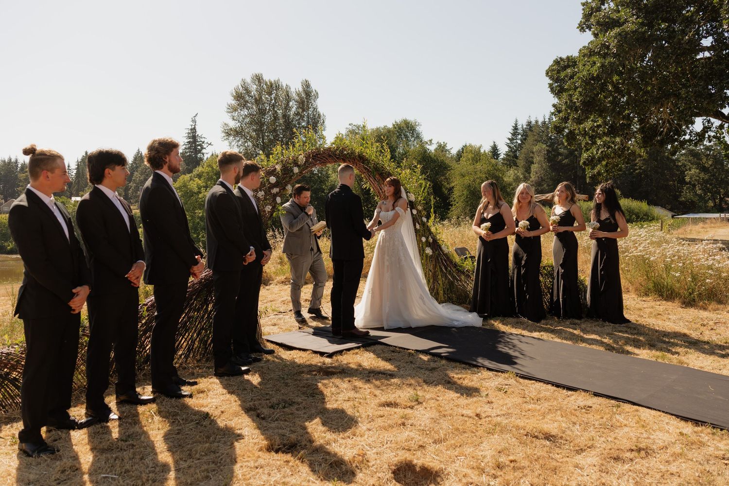 Outdoor wedding ceremony with bride, groom, officiant, groomsmen in black suits, and bridesmaids in black dresses.
