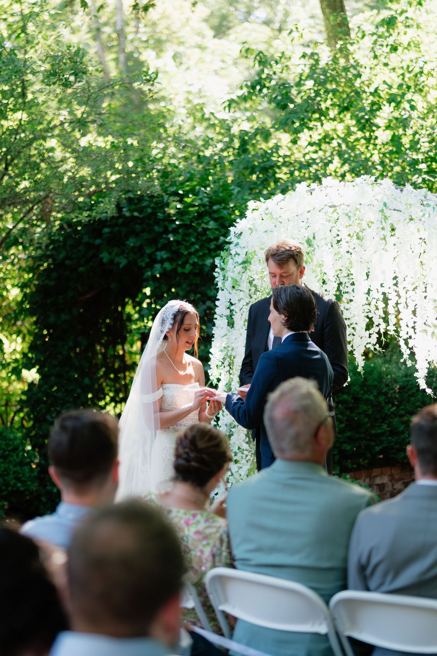 Bride placing ring on groom's finger at outdoor garden wedding ceremony under white floral arch with guests.