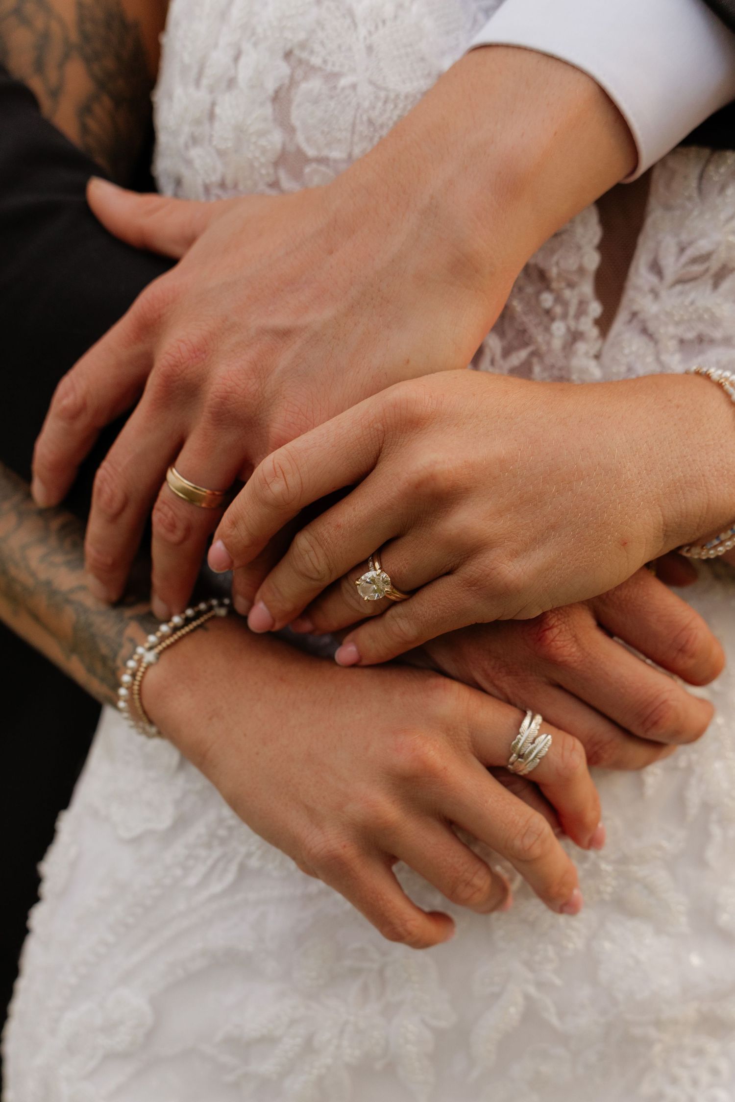 Close-up of newlyweds hands stacked together showing wedding rings, with lace bridal gown and tattooed groom sleeve visible.