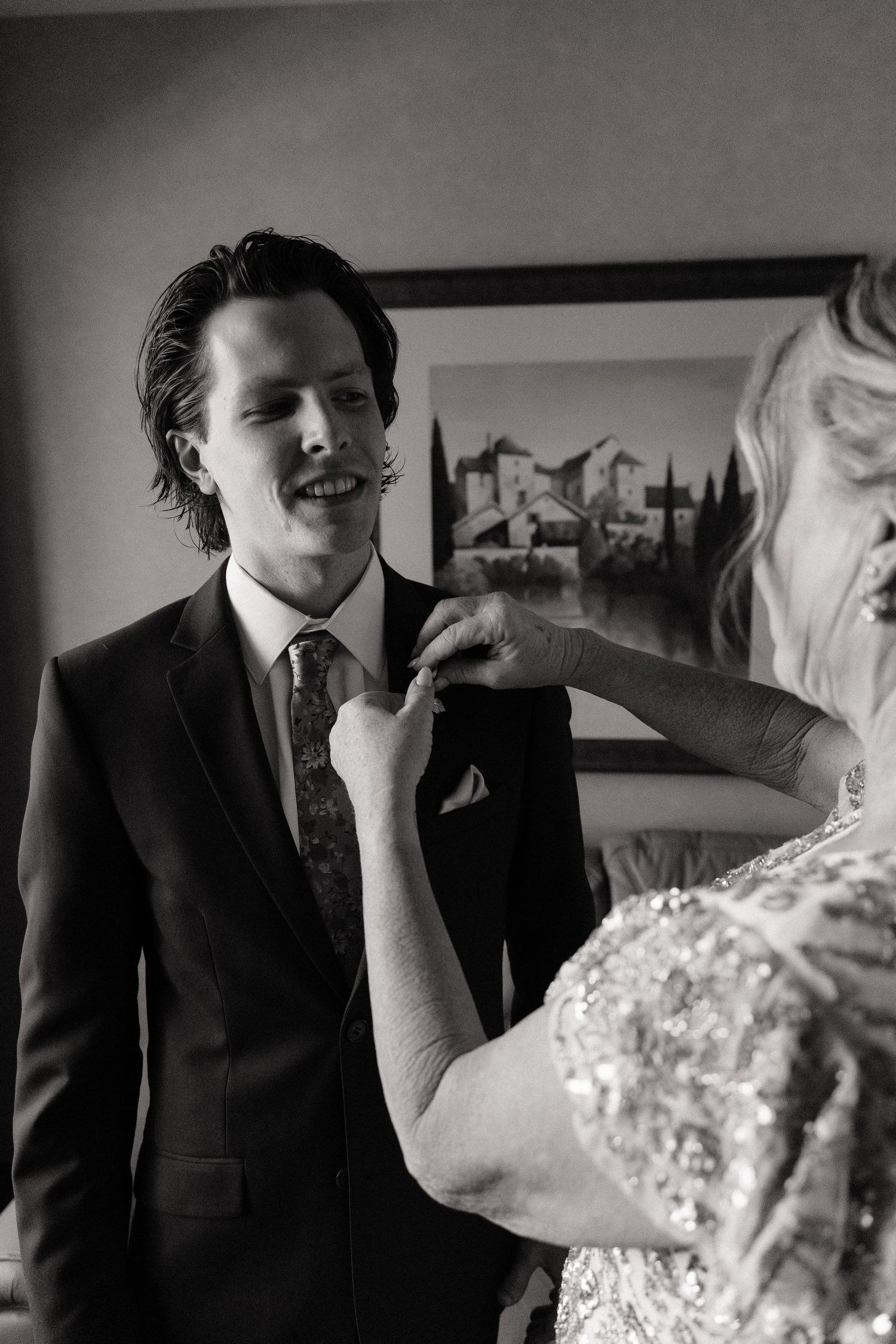 Black and white photo of groom smiling as woman adjusts his boutonniere before wedding ceremony.