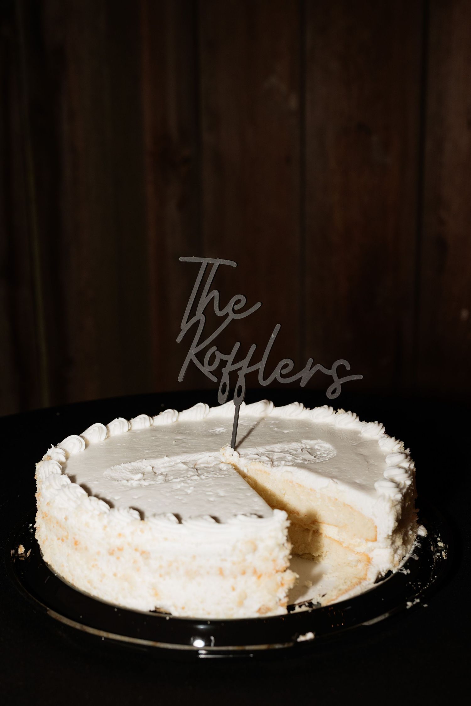 White wedding cake with The Kofflers cake topper, partially sliced, on dark wooden background.