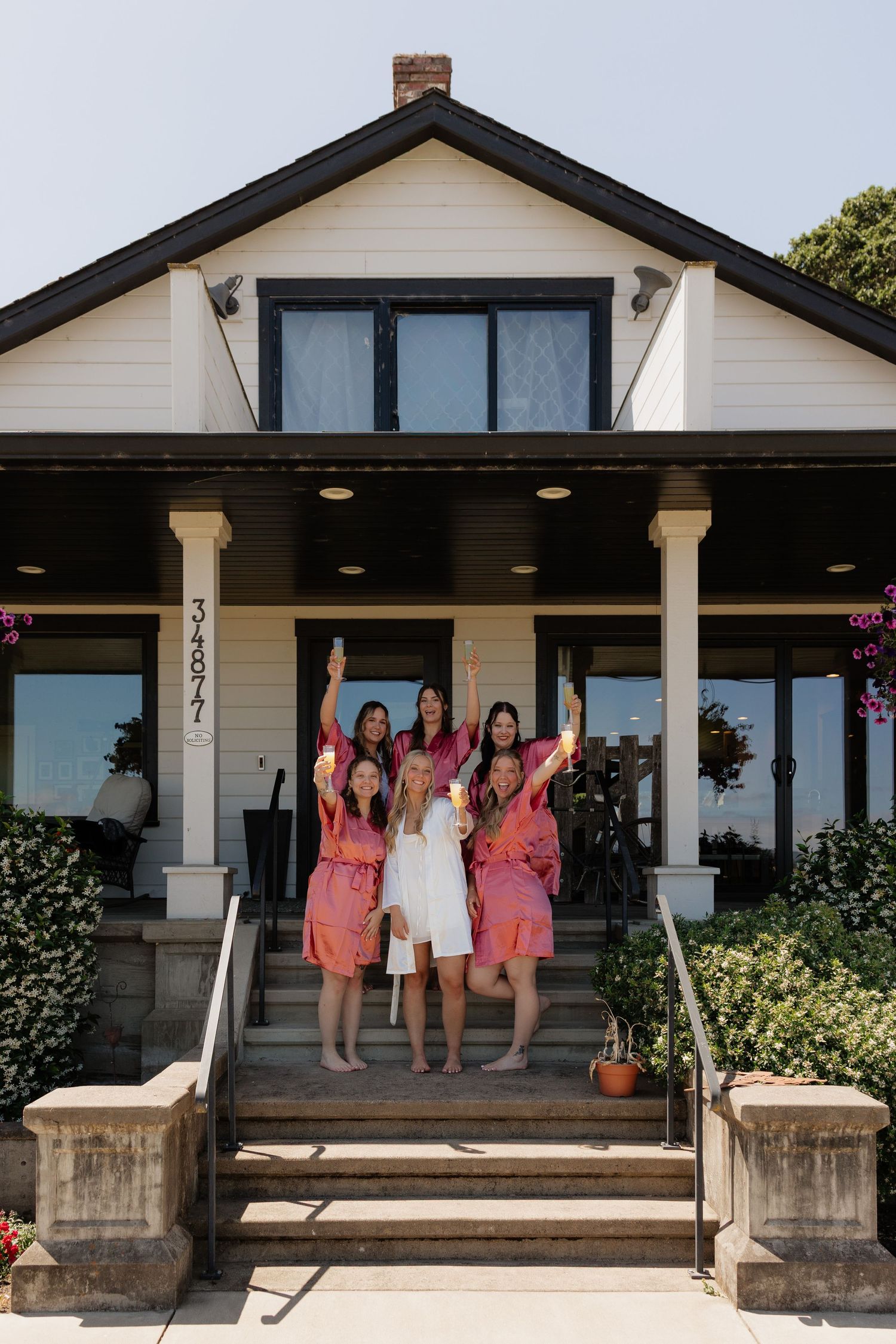 Bride in white robe celebrates with bridesmaids in pink robes on porch steps of white farmhouse.