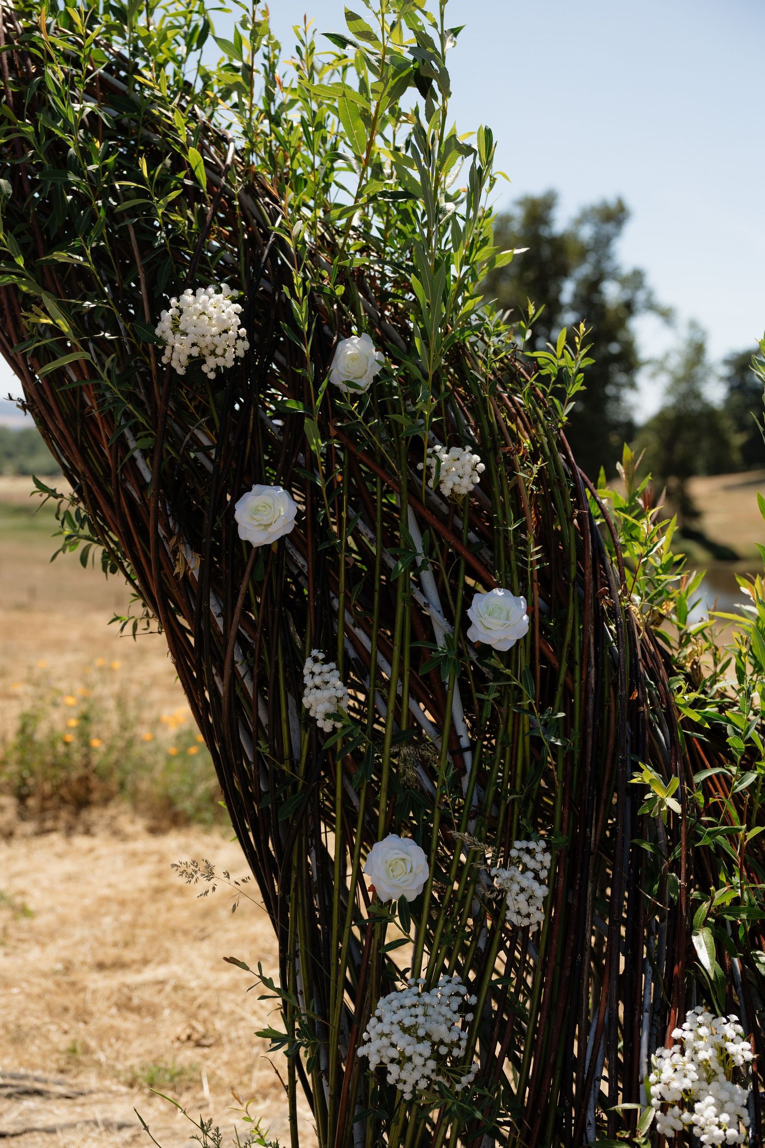 Rustic woven branch arch decorated with white roses and greenery outdoors under a clear blue sky.