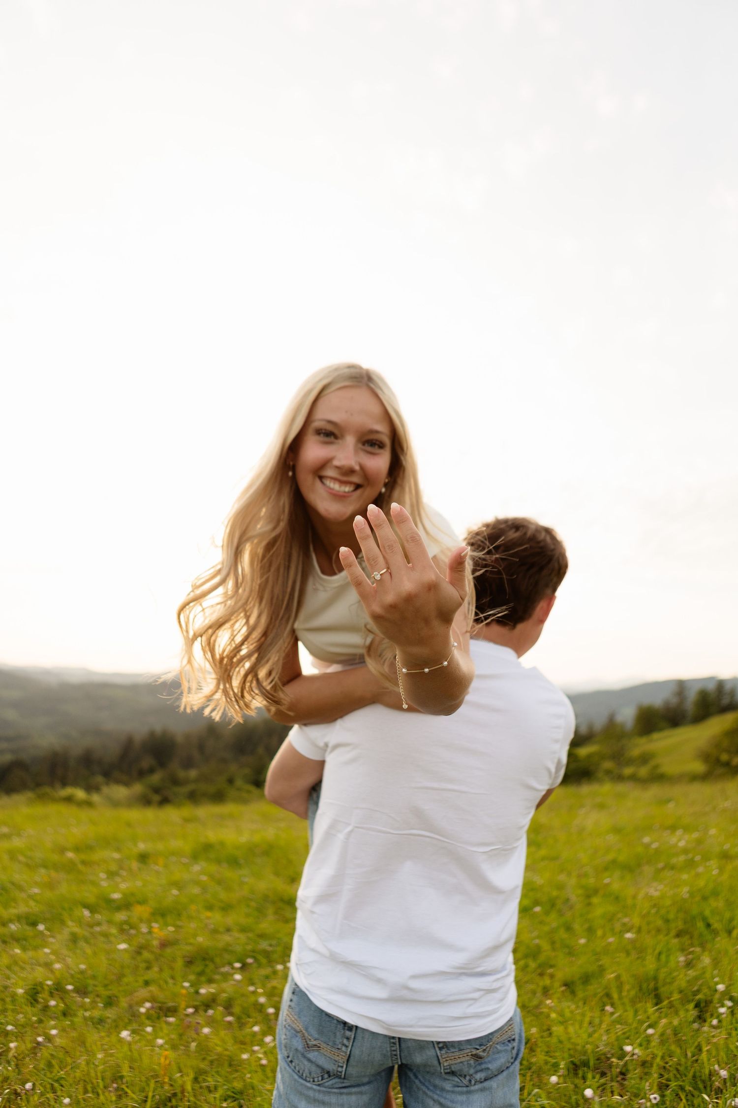 Man lifting smiling blonde woman in green meadow as she shows off engagement ring at sunset.