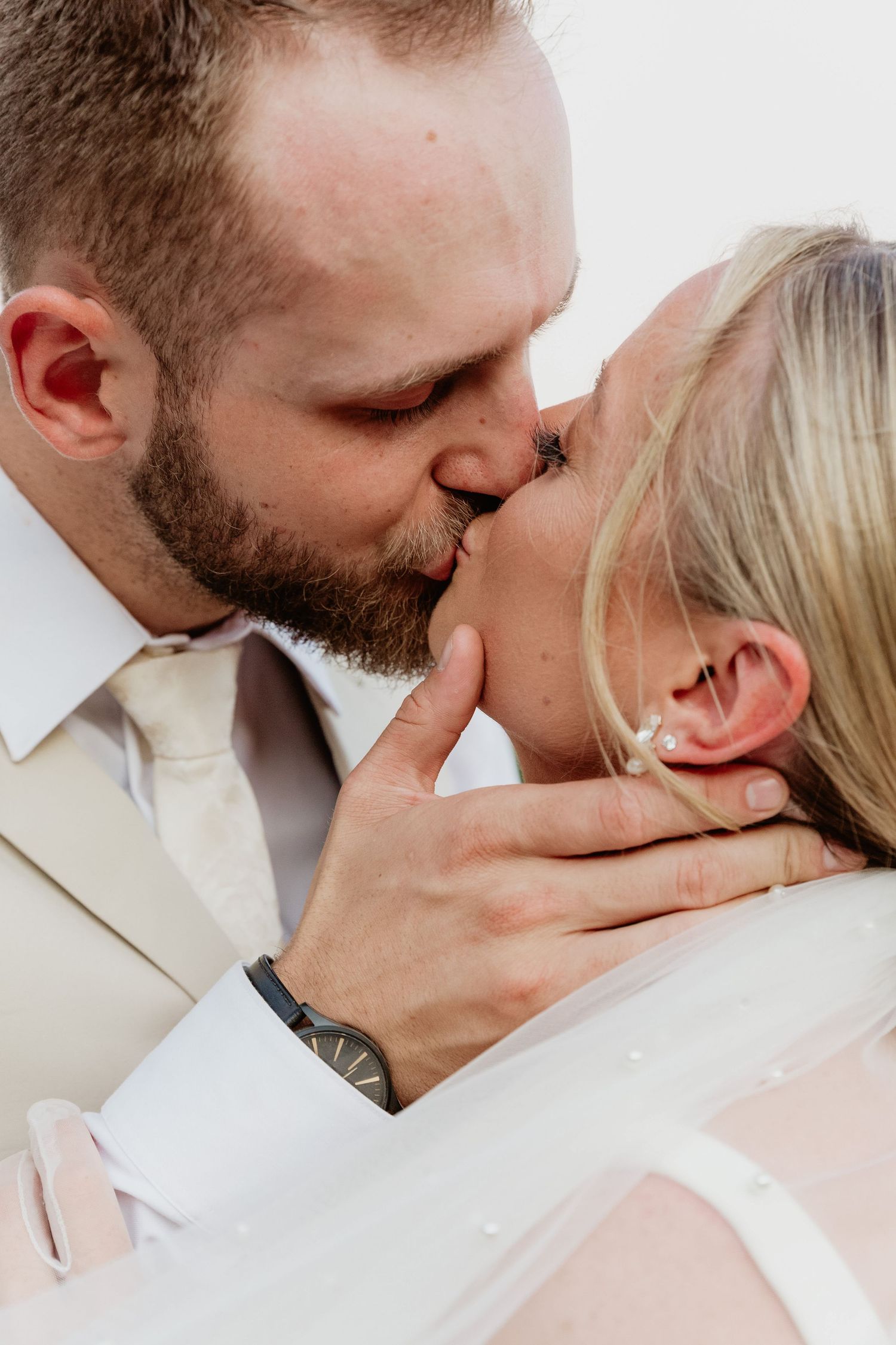 A couple shares a romantic kiss, with the groom in a white suit and watch tenderly holding the bride's face.