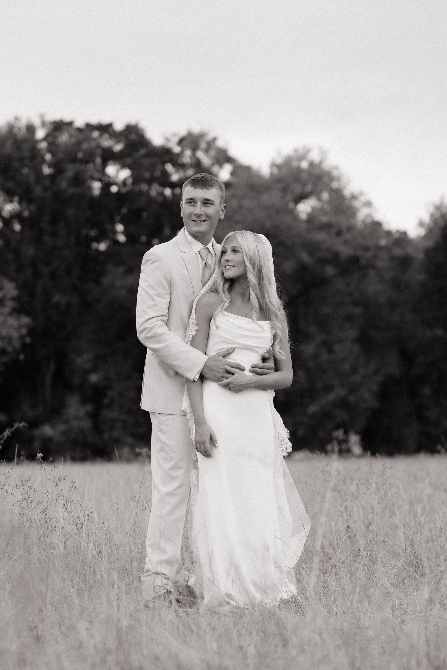 Black and white photo of a couple in formal wear embracing in a field with trees in the background.