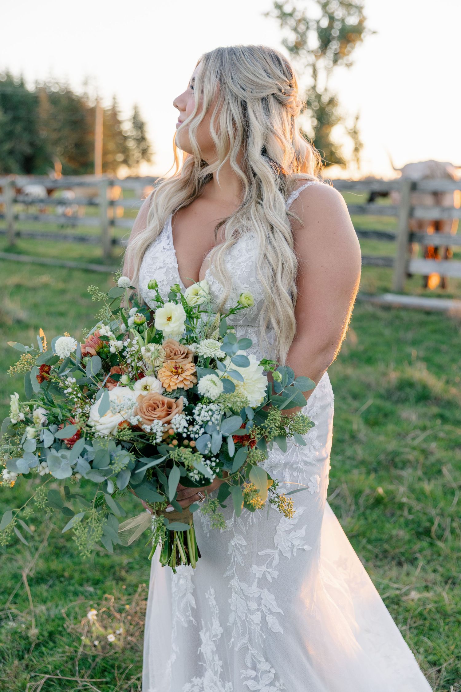 A beautiful wedding bouquet with white flowers and eucalyptus is held by someone wearing a lace wedding dress in a field.