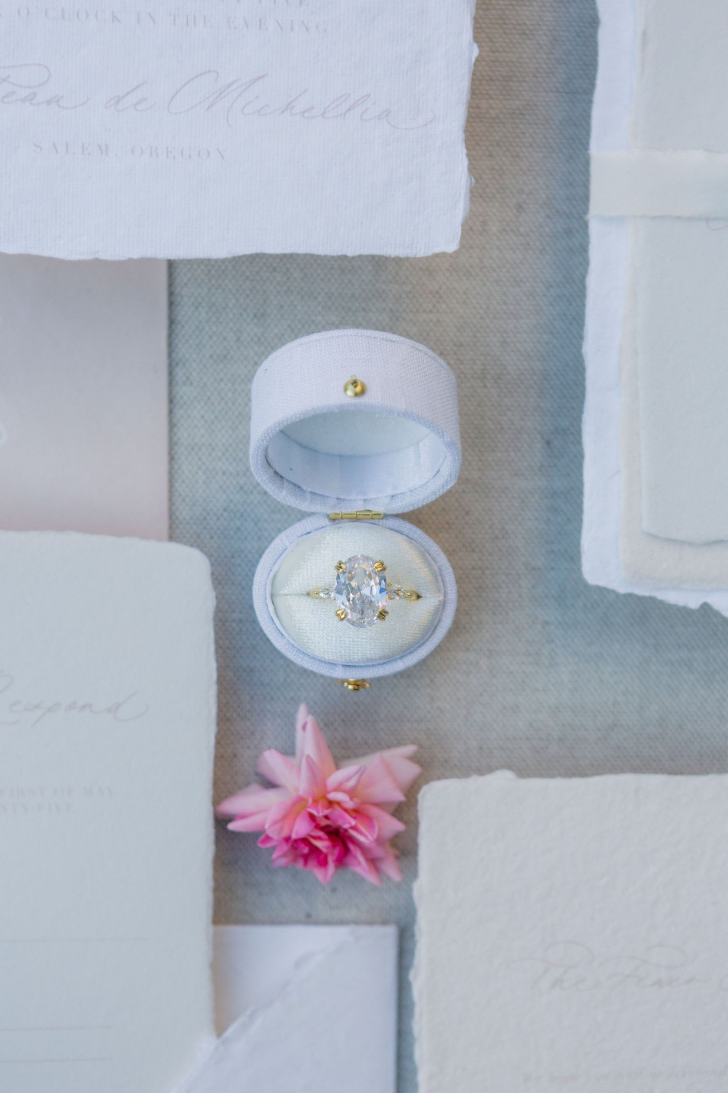 Diamond engagement ring in white velvet box with pink carnation flower on light gray background with paper details.