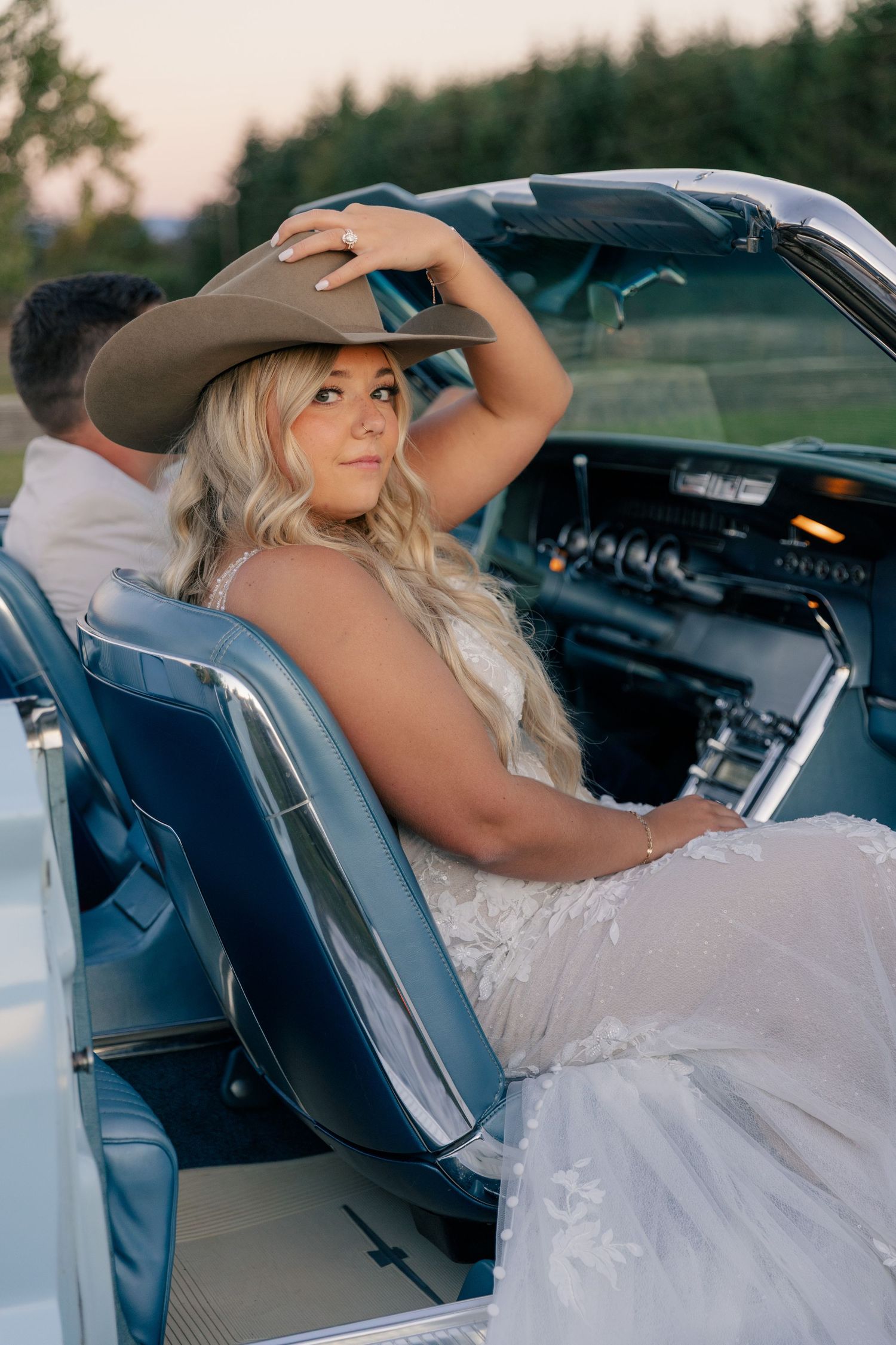 Someone wearing a white wedding dress and cowboy hat sits in a classic convertible car at sunset.