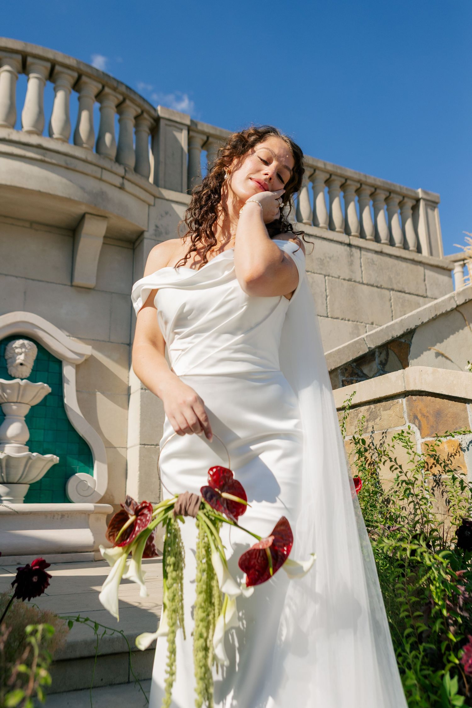 A bride in a white wedding dress poses gracefully along an ornate balustrade with red roses in the garden.
