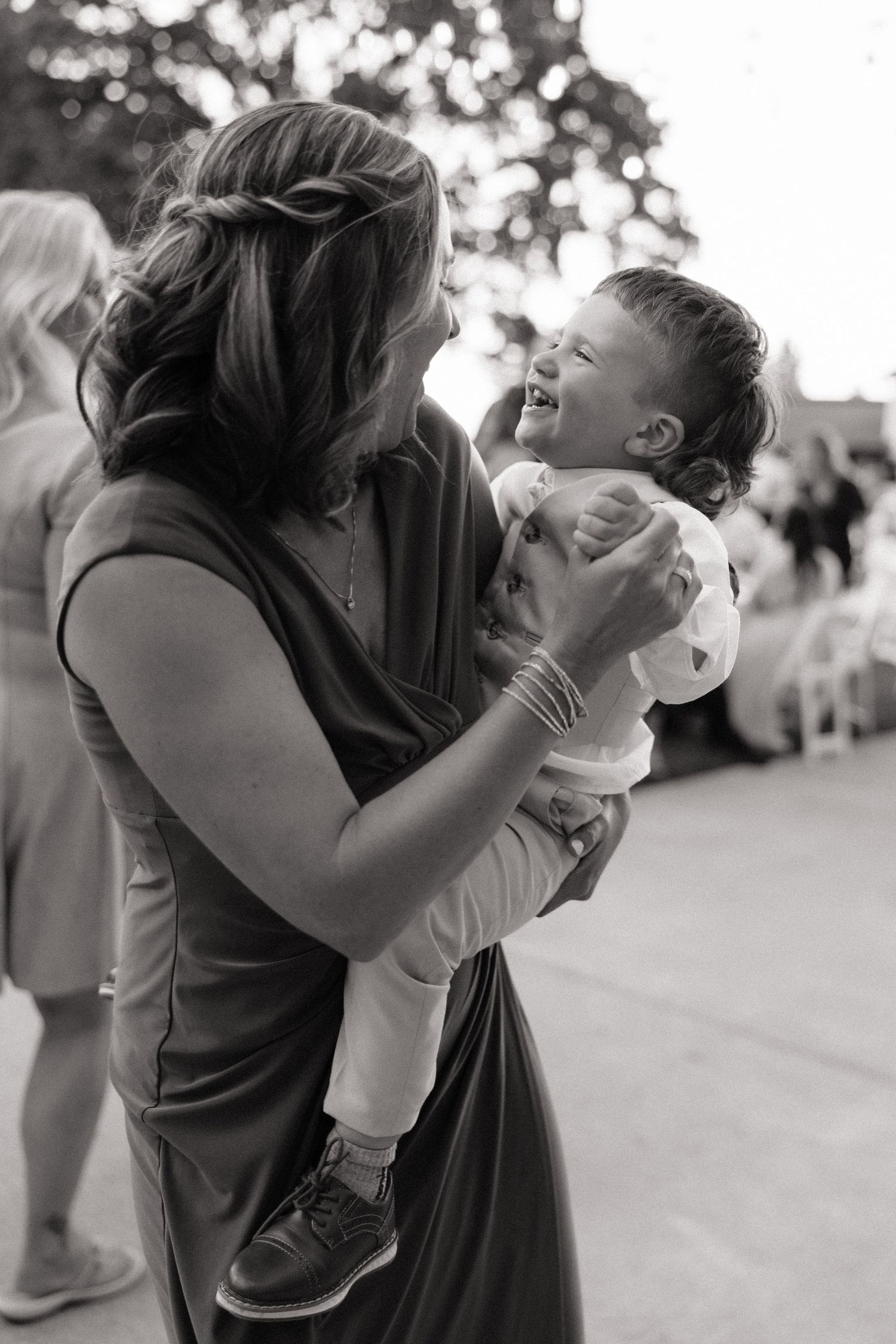 Black and white photo of bridesmaid joyfully holding laughing baby at outdoor wedding reception.