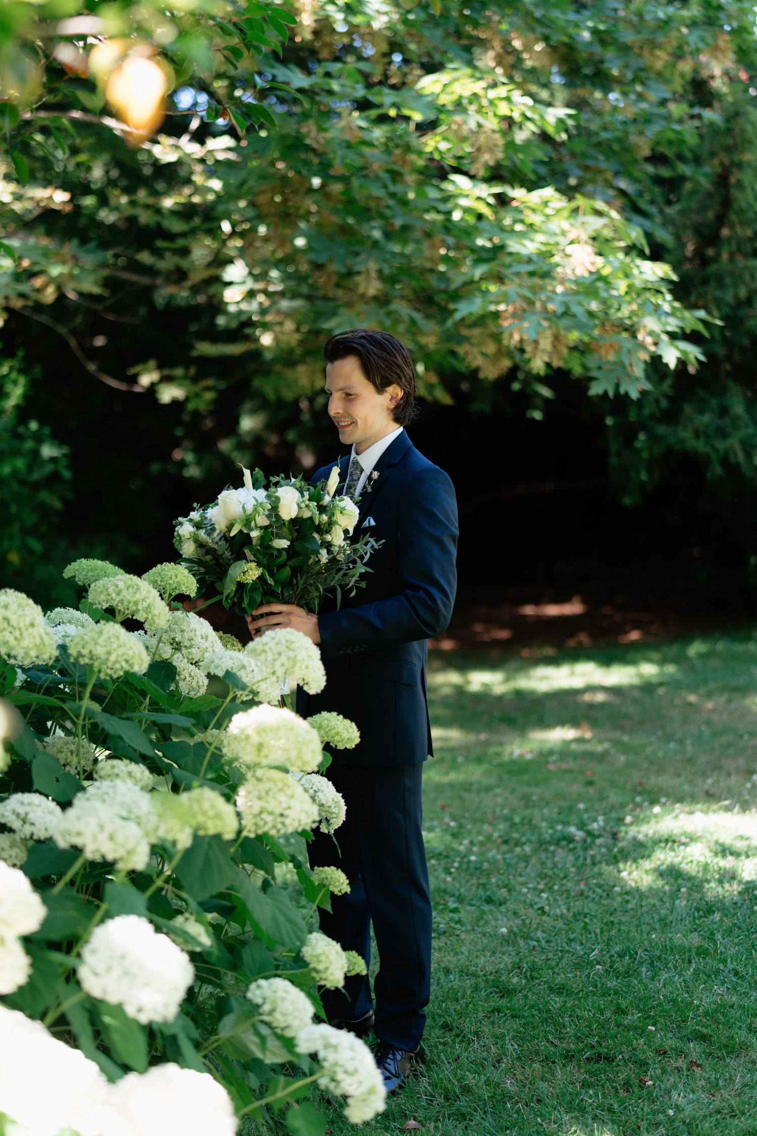 Smiling groom in navy suit holding white floral bouquet among blooming hydrangeas in garden.