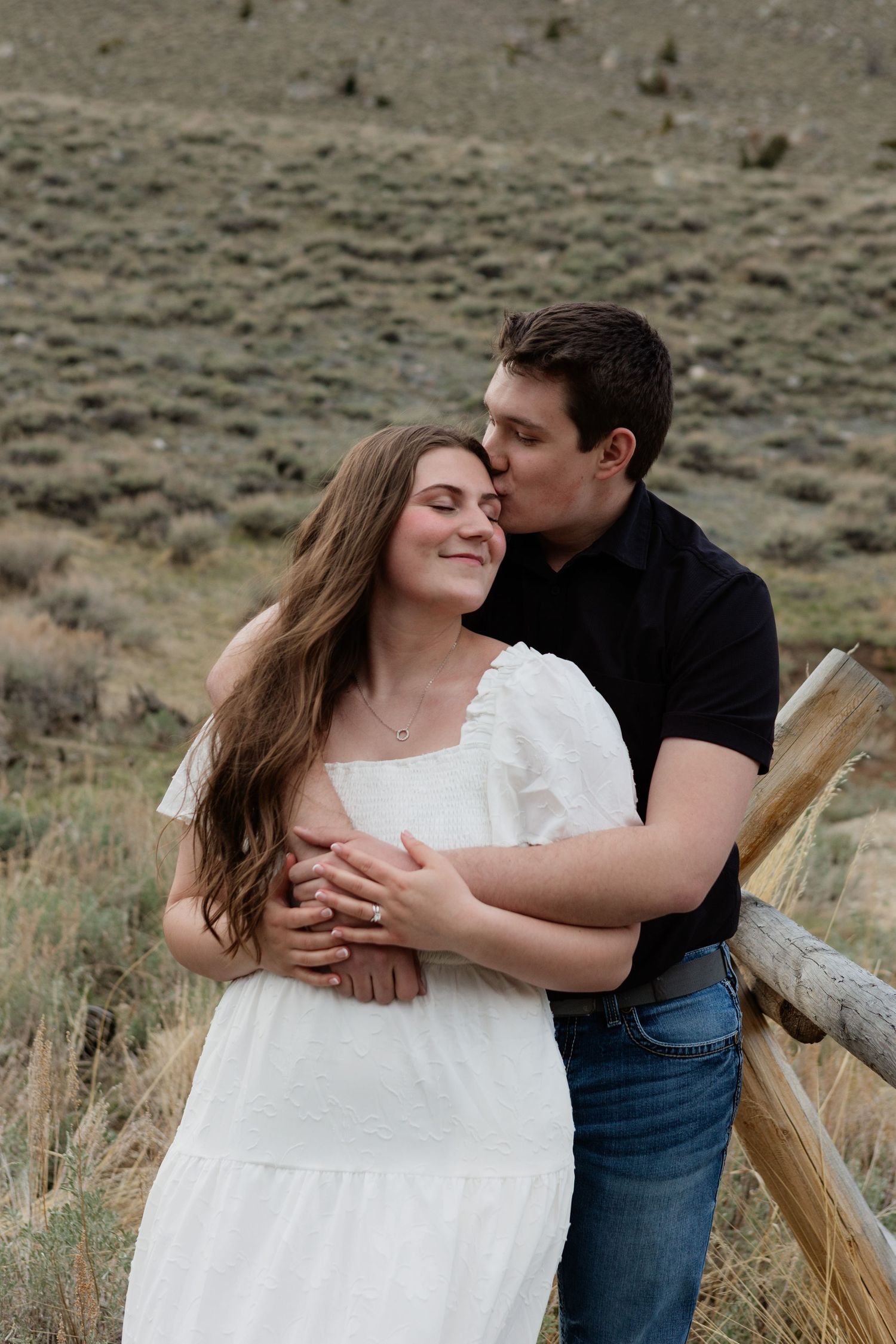 A couple shares a tender moment outdoors, man in black shirt kissing woman in white dress near a wooden fence.