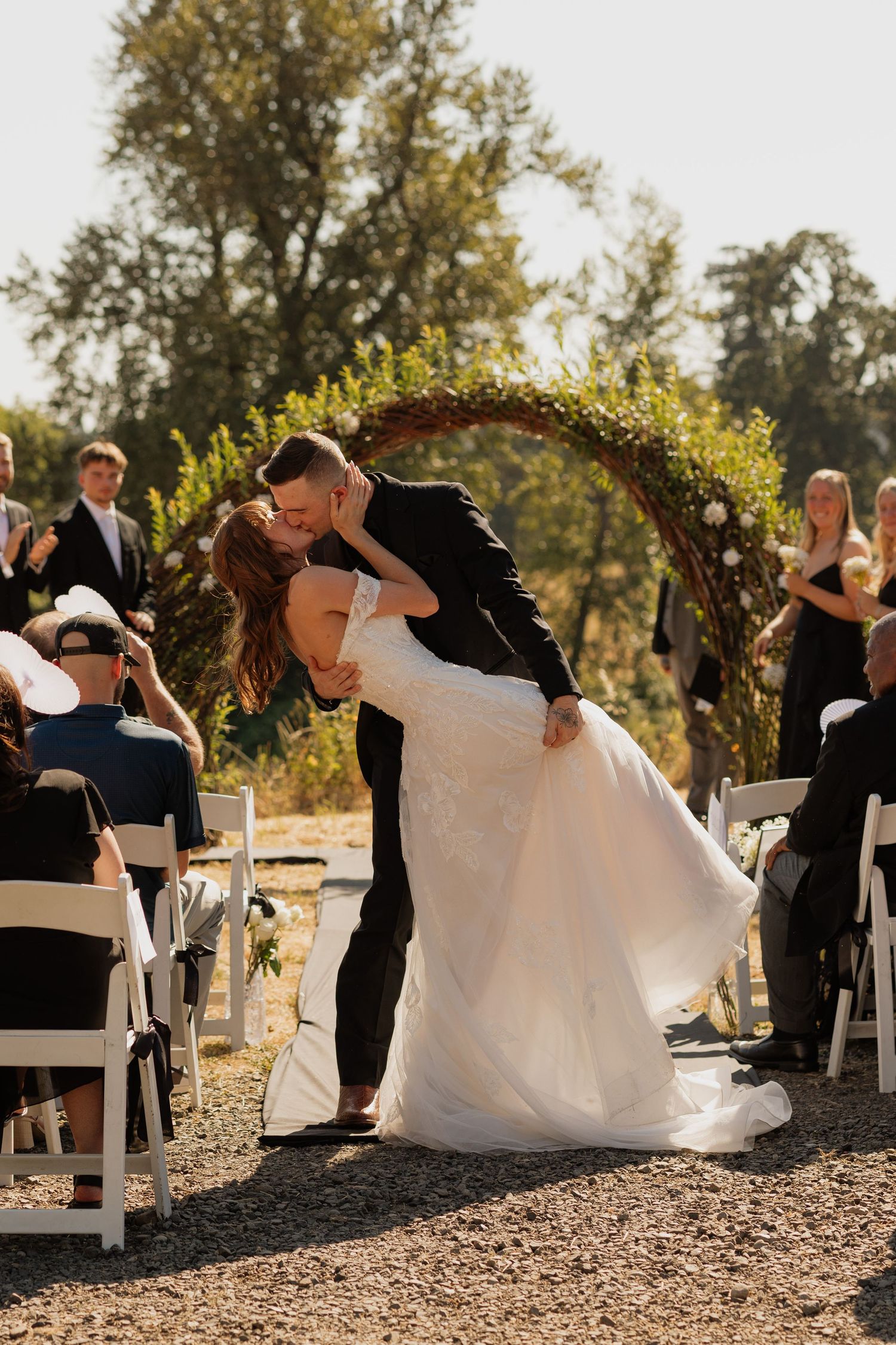Bride and groom share a romantic dip kiss at outdoor wedding ceremony beneath a lush circular floral arch.
