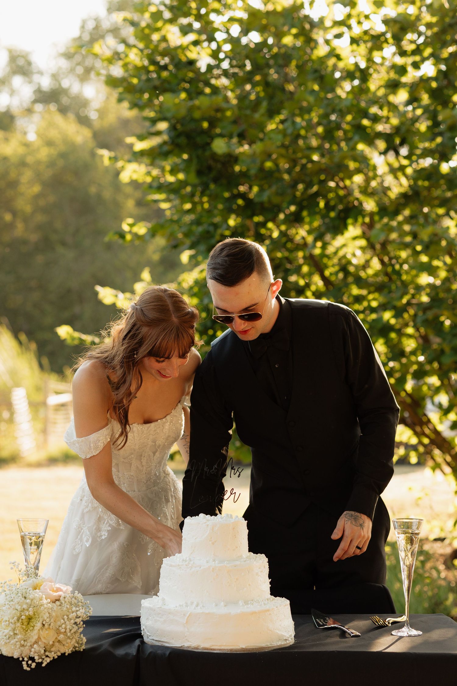 Bride and groom cutting a white three-tier wedding cake outdoors surrounded by lush green trees.