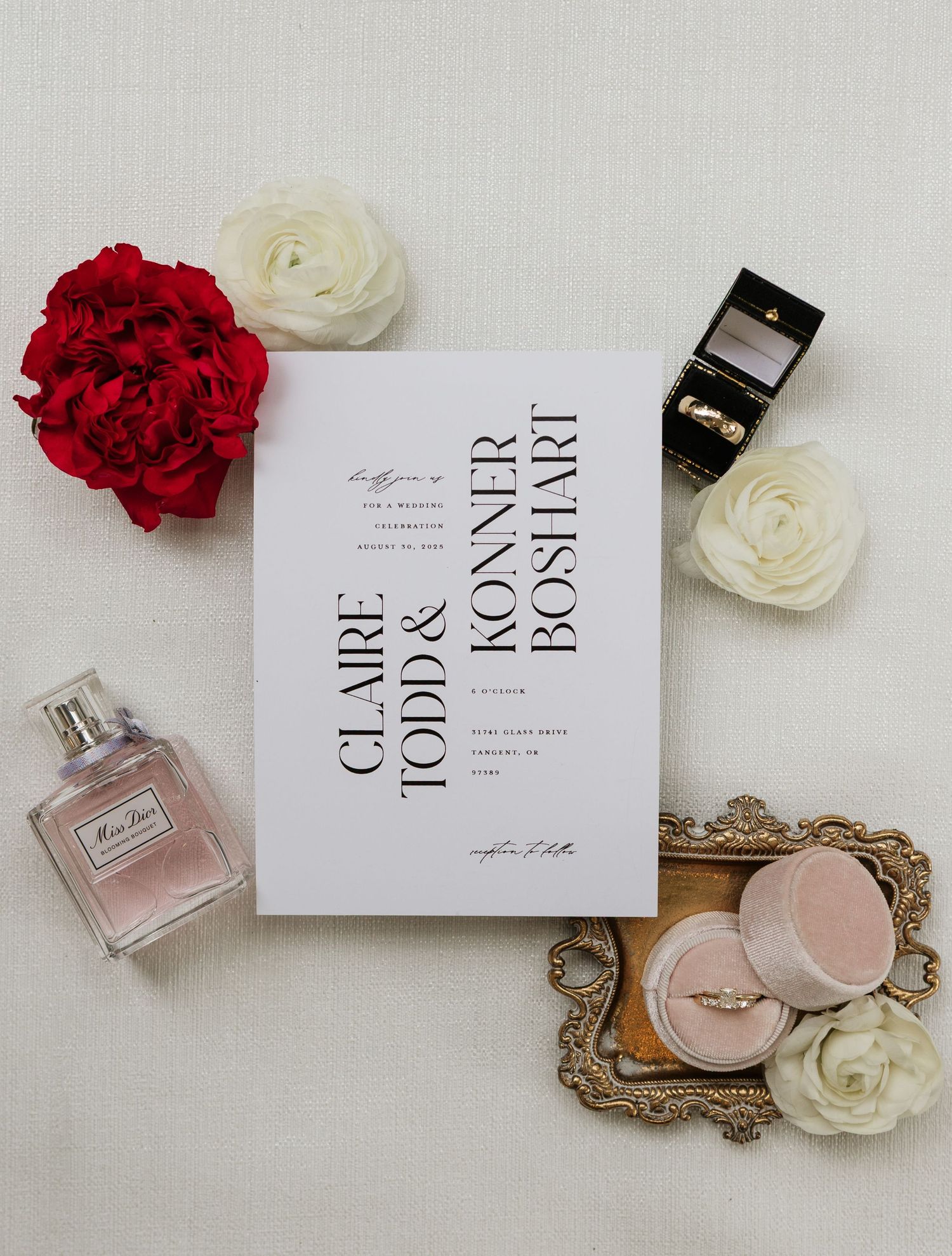 Elegant wedding invitation for Claire Todd and Konner Boshart displayed with roses, a ring box, and Miss Dior perfume.