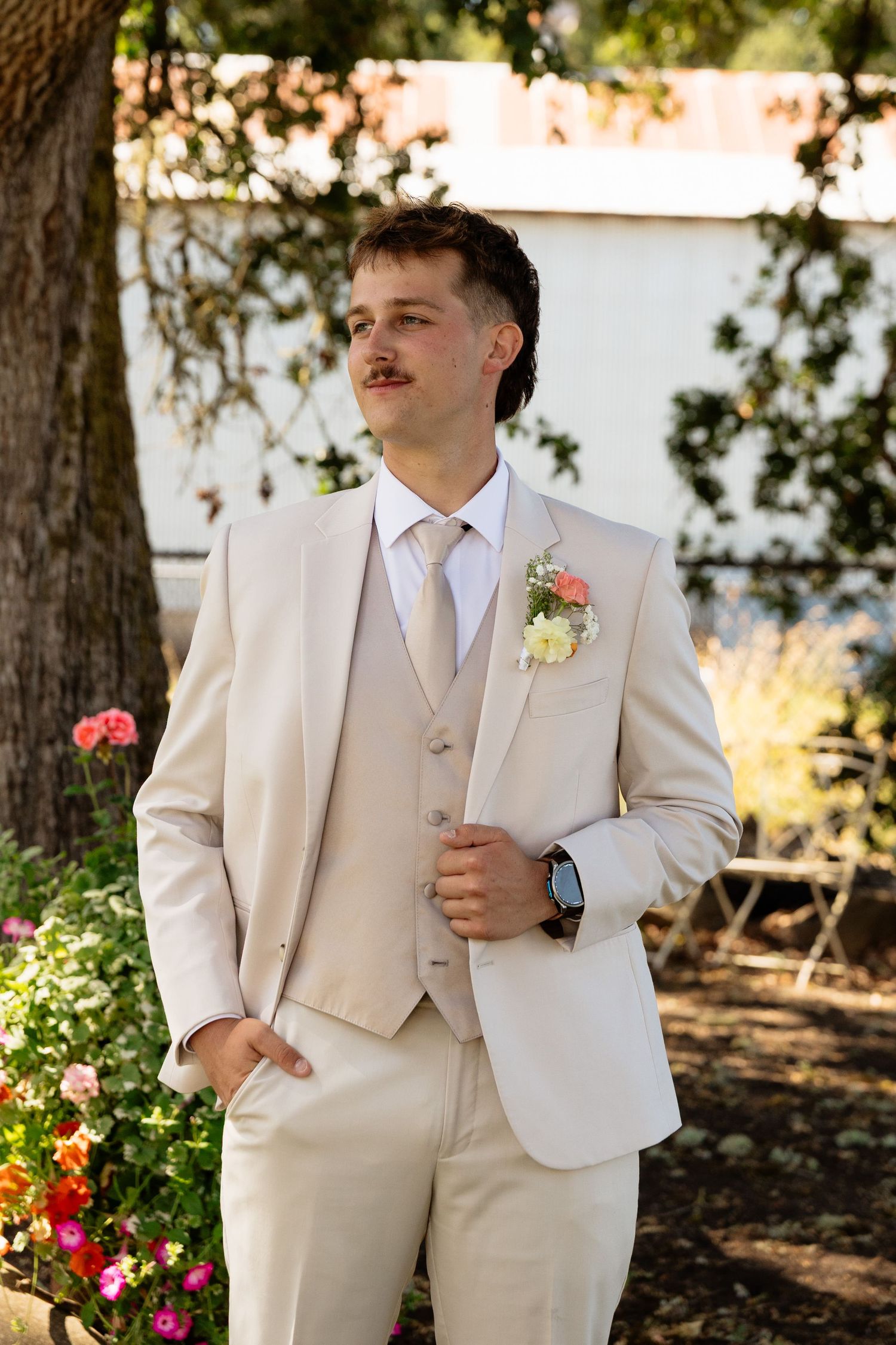 Groom in beige three-piece suit with floral boutonniere standing outdoors among garden flowers on wedding day.