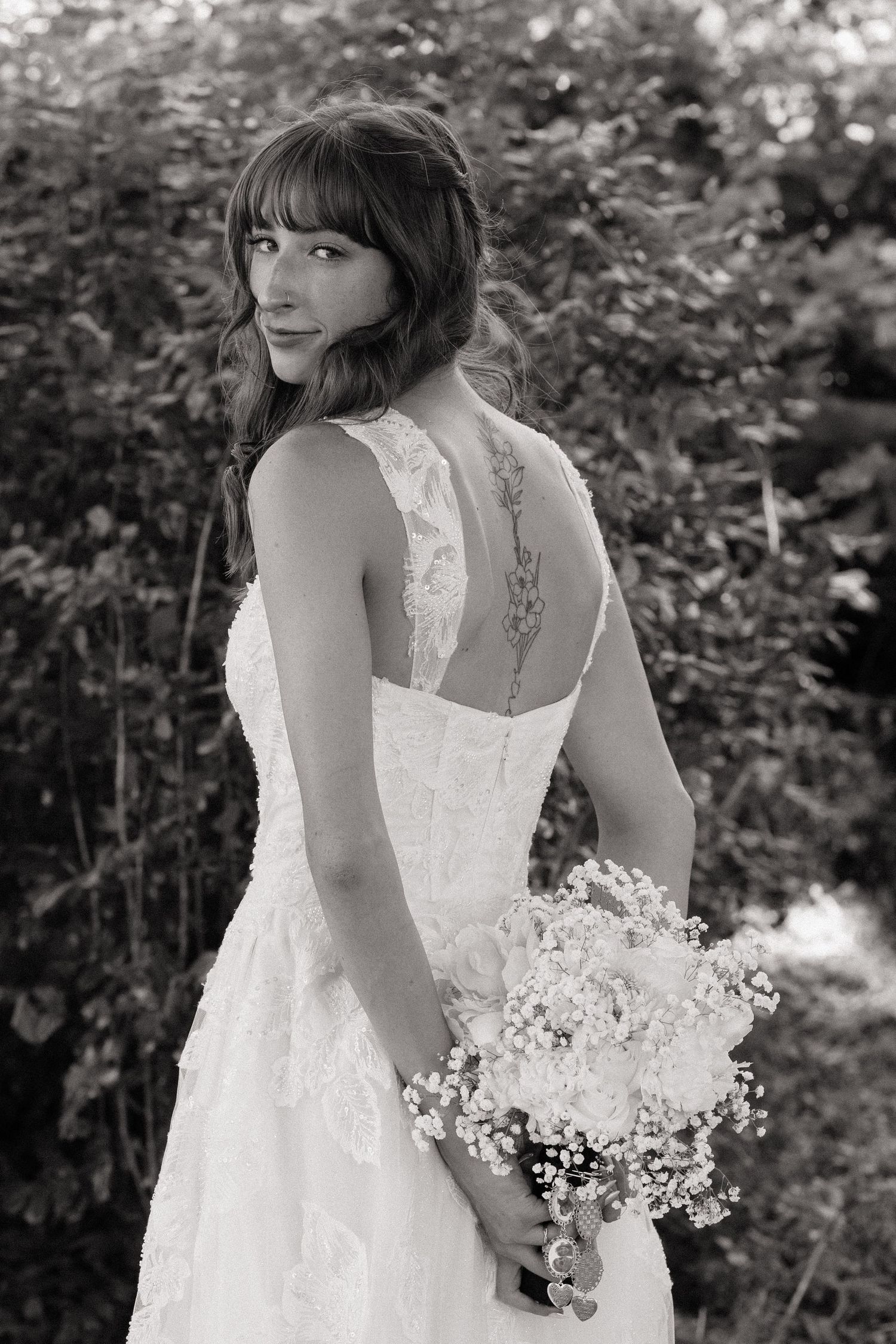 Bride in lace wedding gown holds bouquet, showing back detail with floral tattoo, black and white photo.