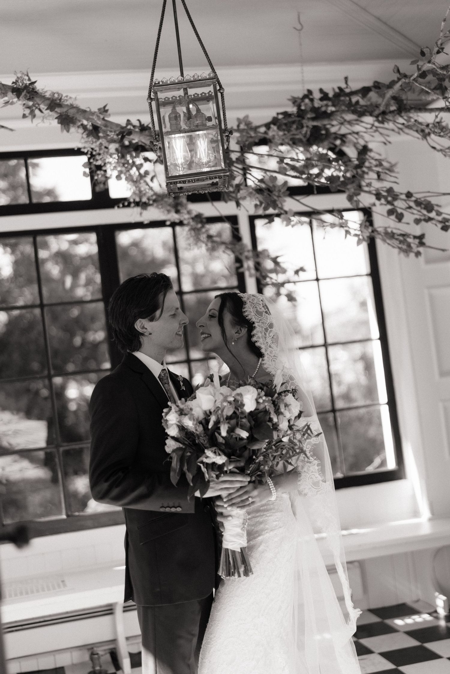 Black and white photo of bride and groom sharing a tender moment under floral garland with hanging lantern.