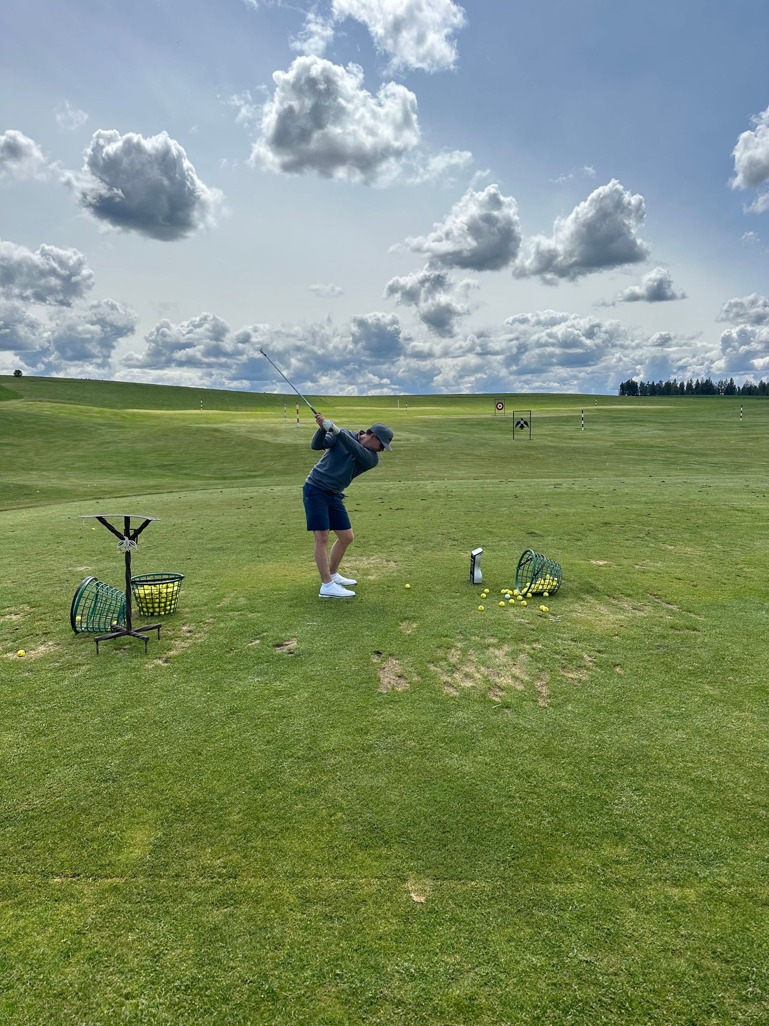 Golfer mid-swing on a lush green driving range under a partly cloudy sky with yellow ball baskets nearby.