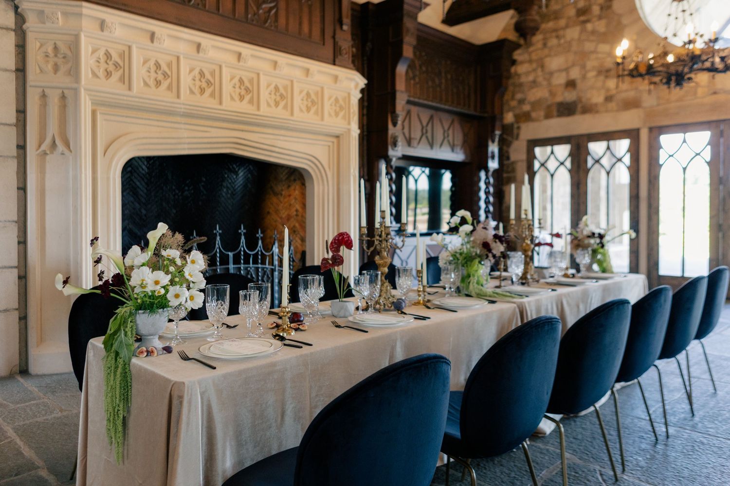 A long banquet table with navy chairs is set elegantly in an ornate white room with chandelier lighting.