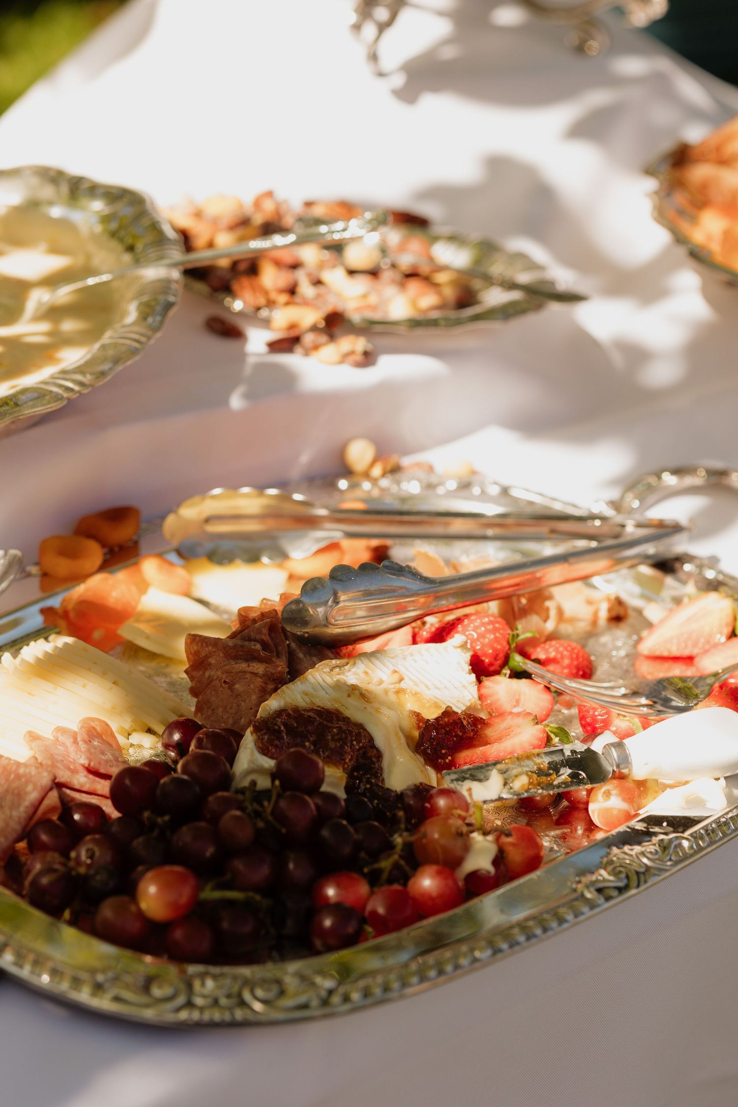 Elegant silver platters with fruit, cheese, and wedding reception appetizers on white tablecloth.