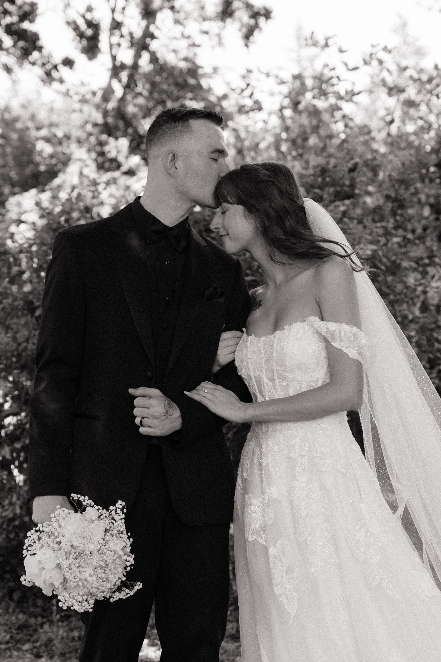 Black and white photo of bride and groom sharing a romantic moment, bride in off-shoulder lace gown holding bouquet.
