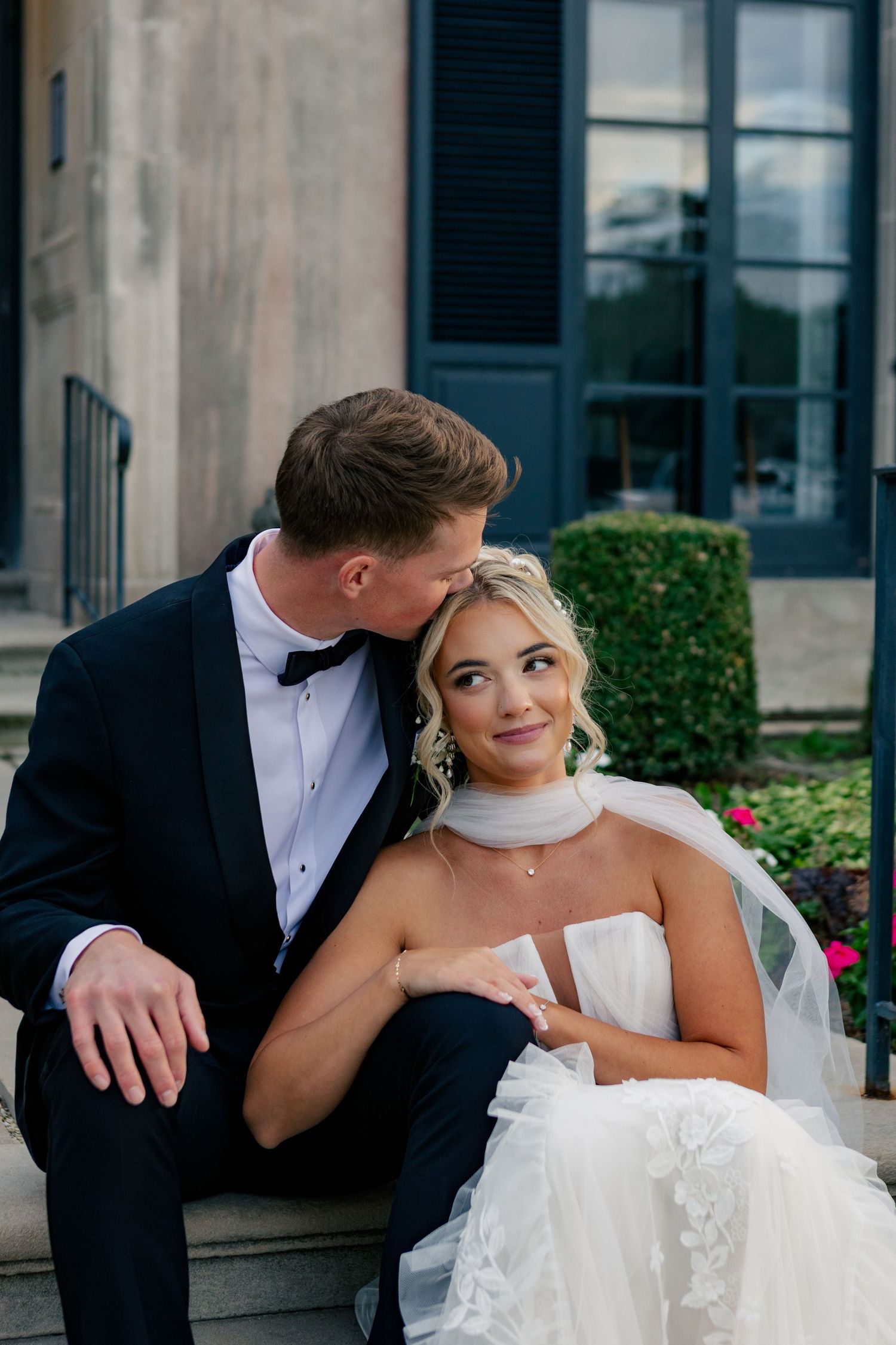Groom in black tuxedo kissing bride in strapless white gown on steps of elegant venue.