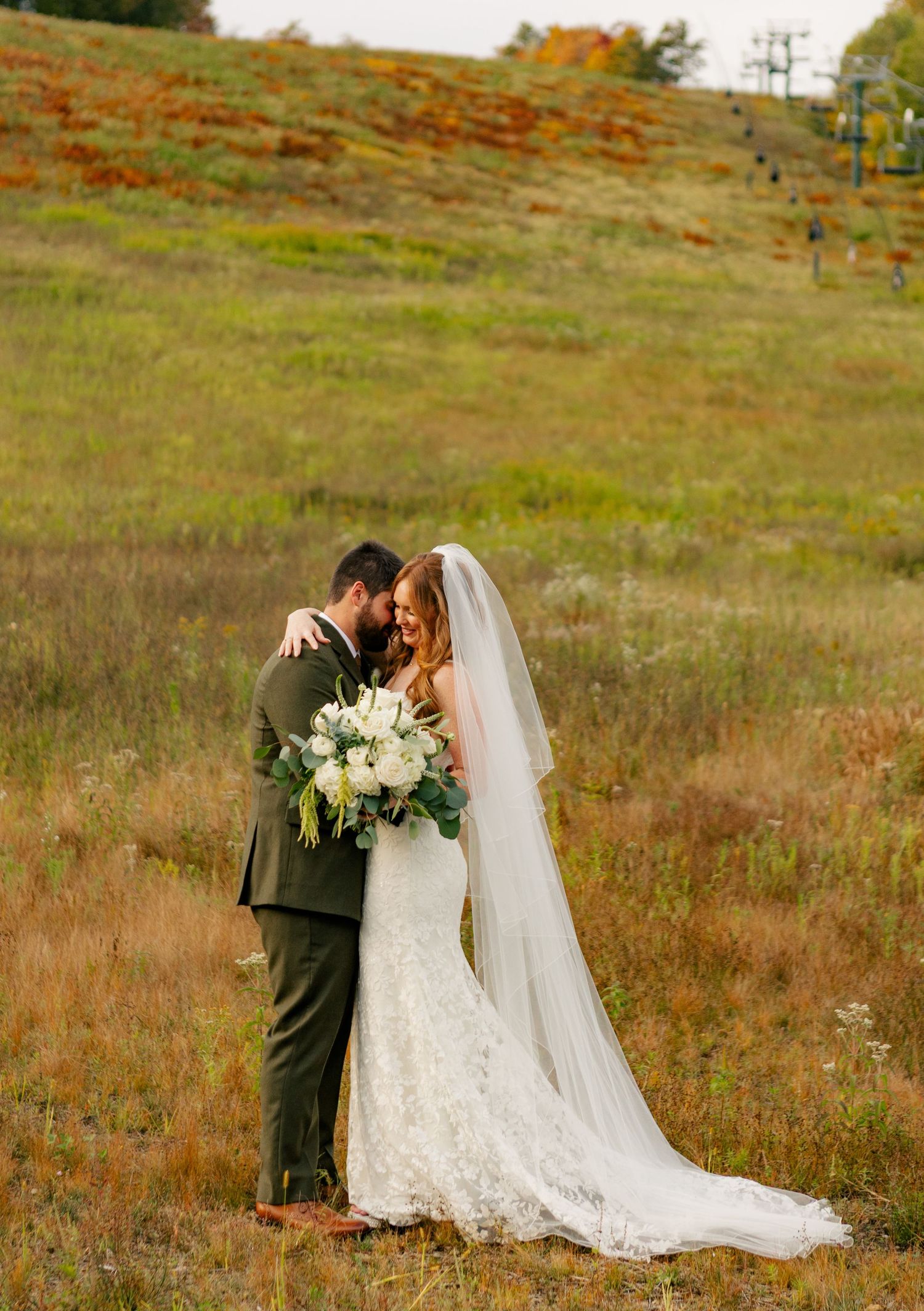 A bride and groom embrace in a golden meadow, her lace gown and veil trailing as she holds a white bouquet.