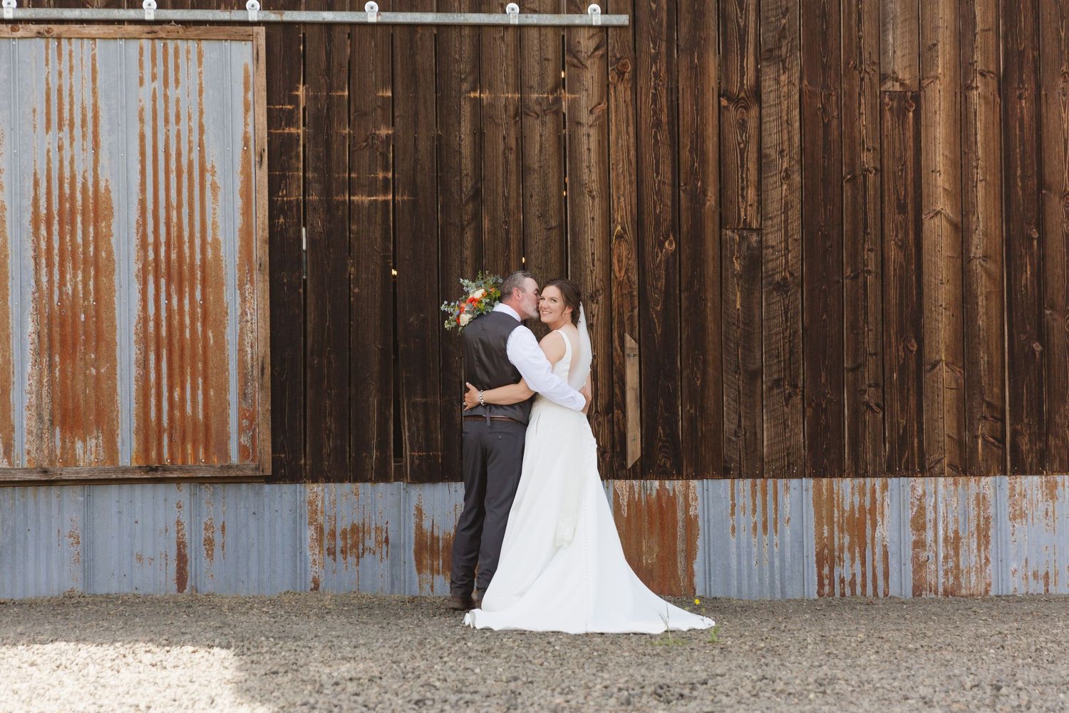 A couple embraces in formal wedding attire against a rustic barn wall with corrugated metal and wood siding.