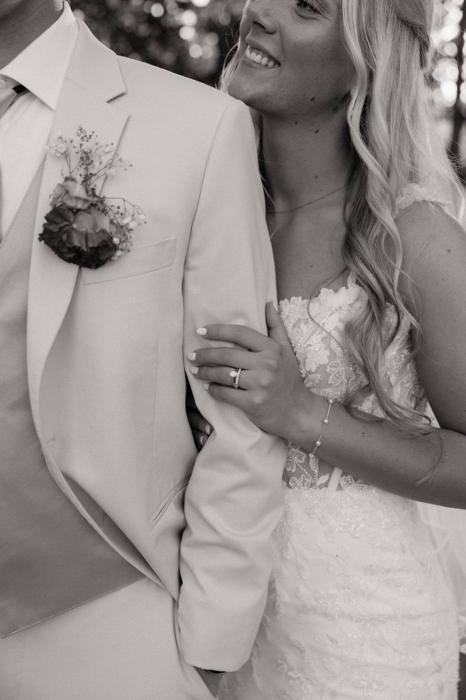 Black and white photo of smiling bride in lace dress holding grooms beige suit jacket arm.