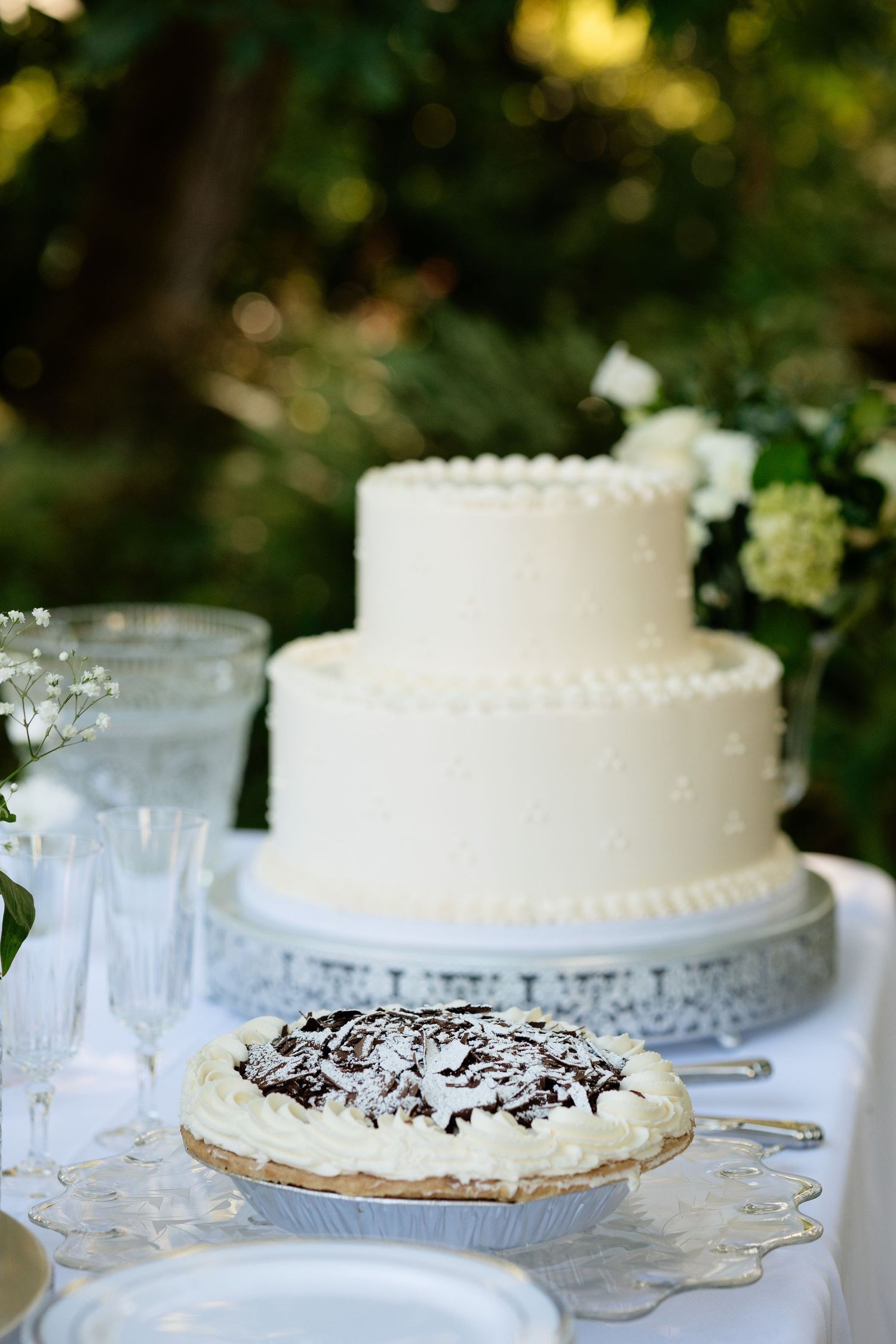 Elegant two-tier white wedding cake with polka dot frosting beside a chocolate coconut pie at an outdoor reception.