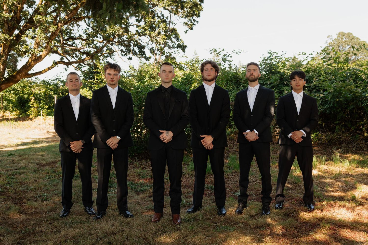 Six groomsmen in matching black suits stand outdoors near green trees for a classic wedding party photo.