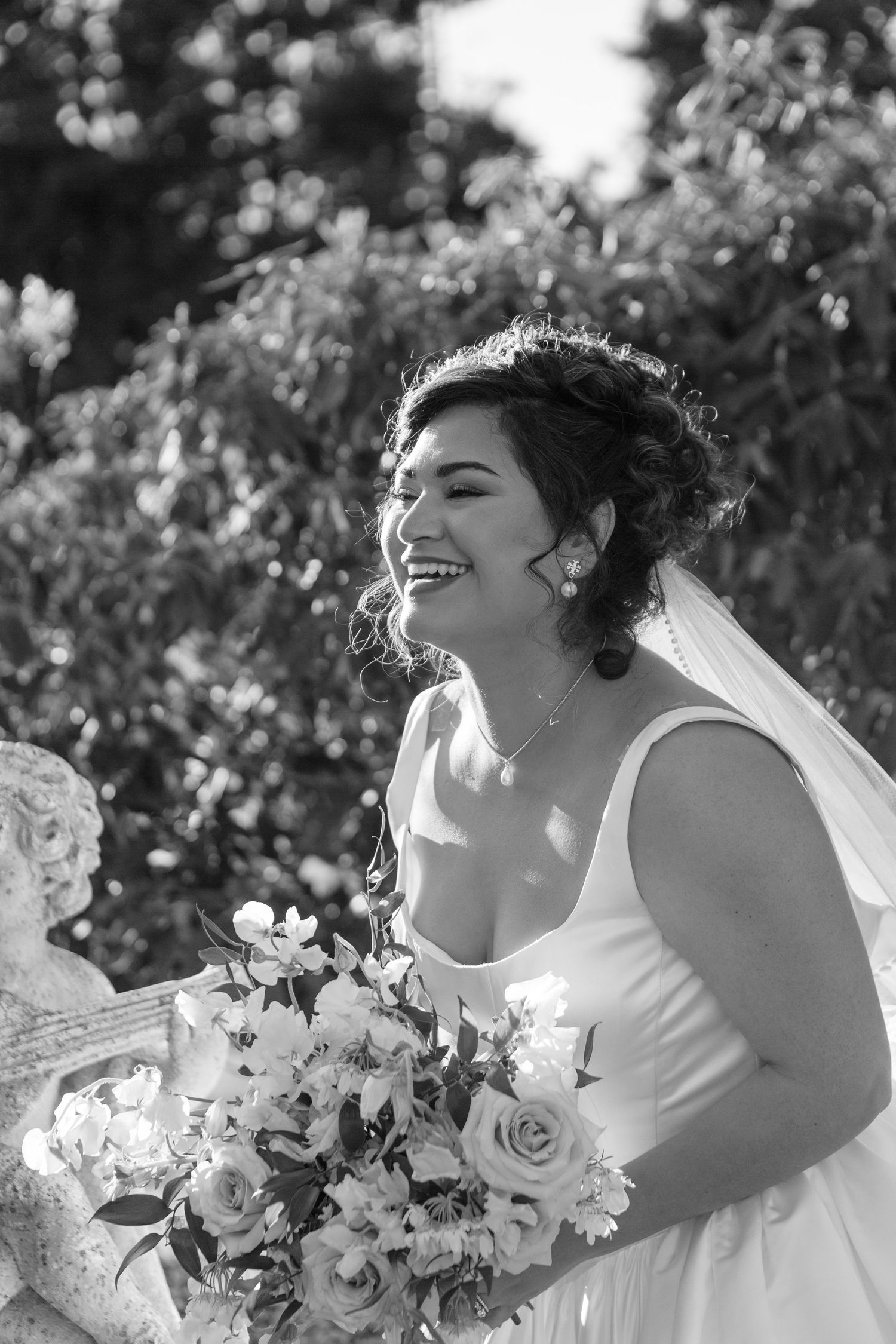 A joyful bride in a white dress laughs while holding a beautiful bouquet of roses and daisies in the sunlight.