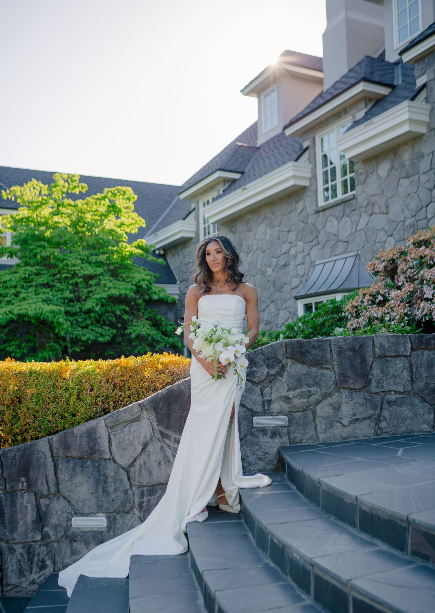 A flowing white wedding dress billows in the breeze against a rustic stone wall and garden backdrop.