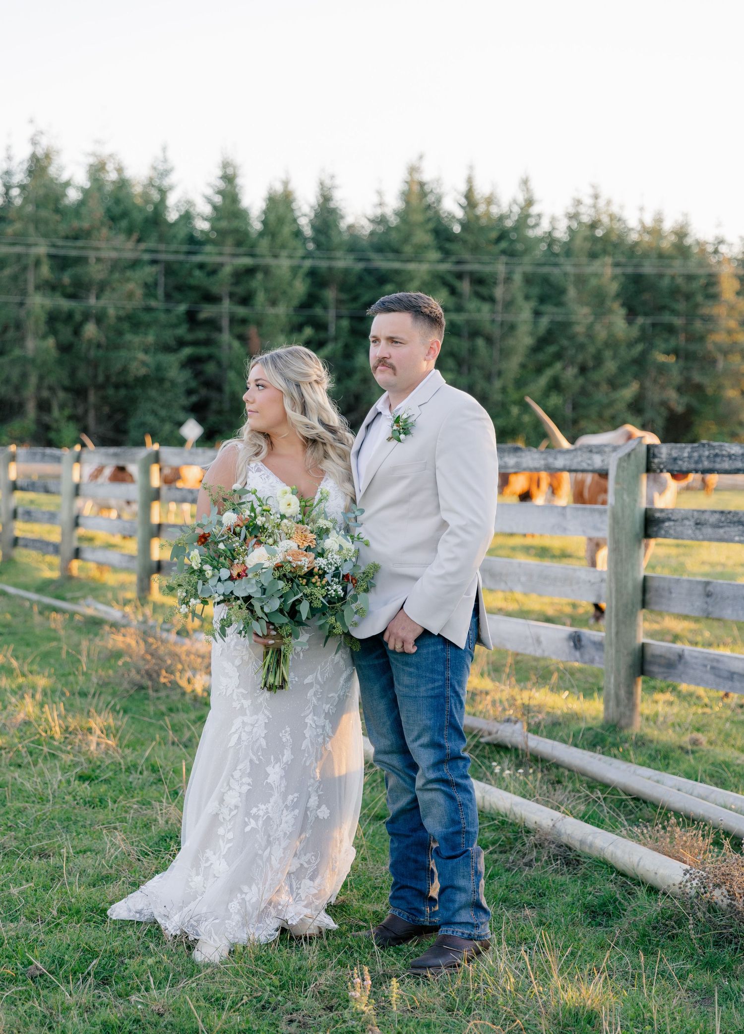 A couple stands by a rustic wooden fence at sunset on a farm with trees and glowing lights in the background.