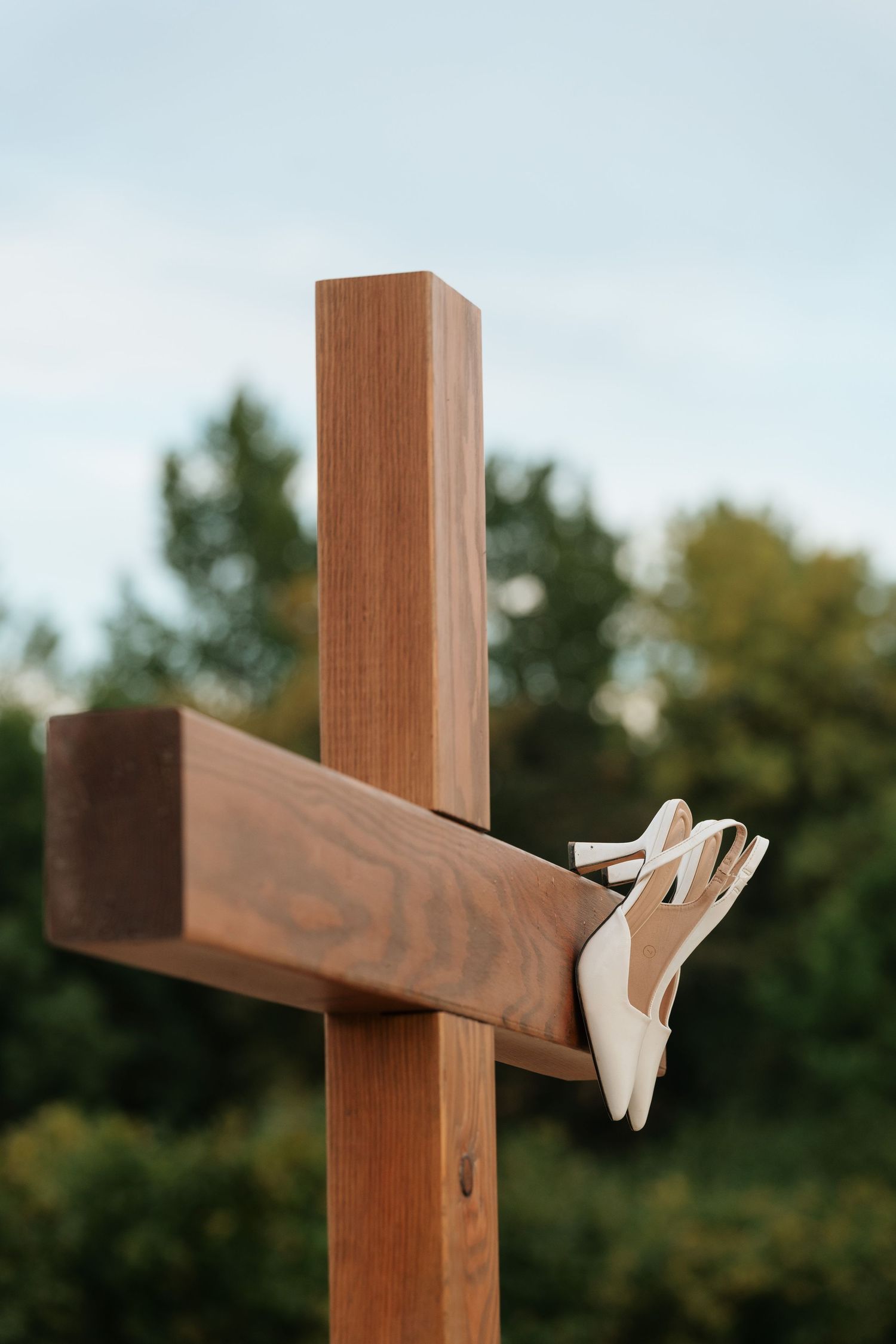 White bridal heels displayed on a rustic wooden cross outdoors with green trees in the background.