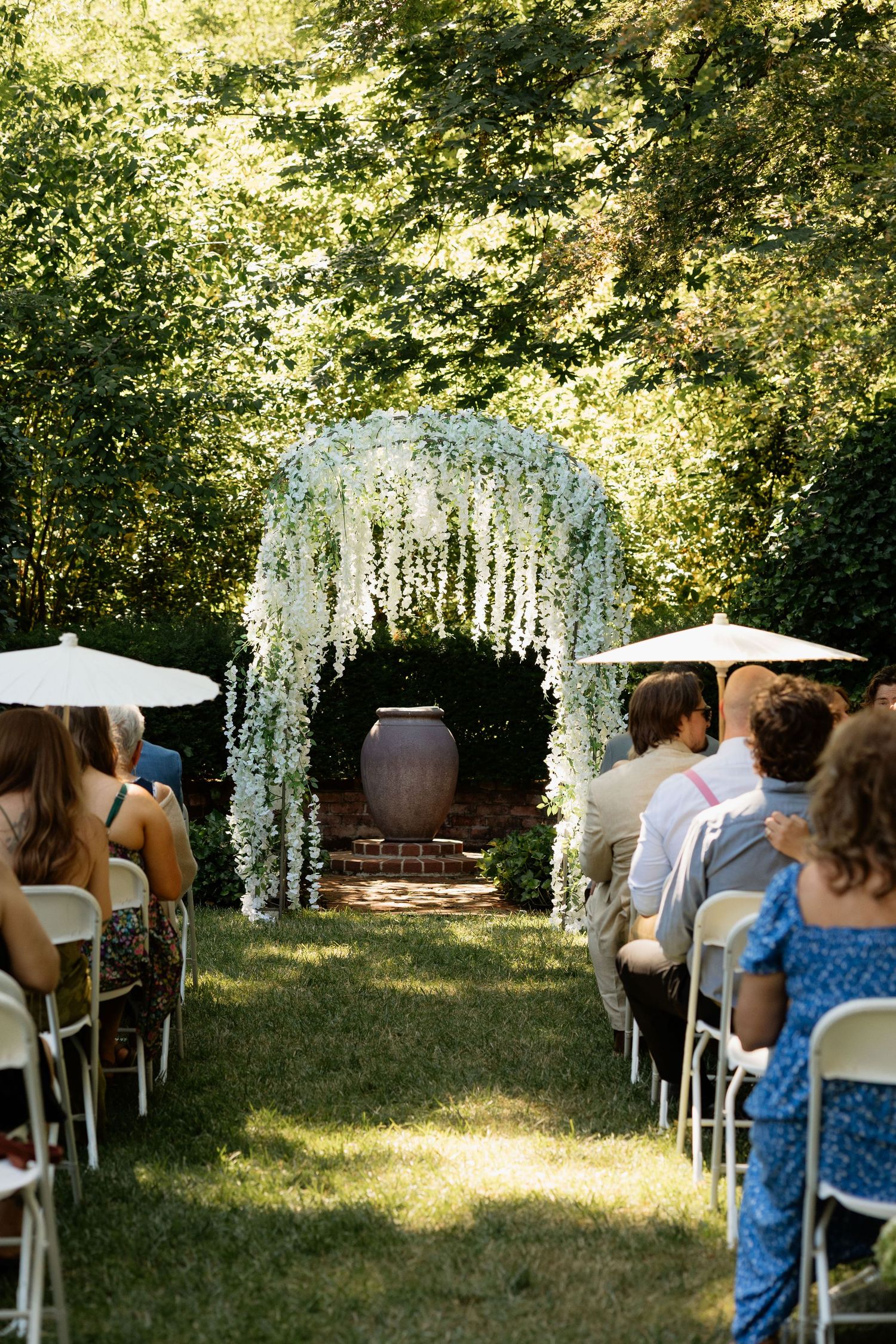 Outdoor wedding ceremony with white floral arch, guests seated on white chairs, lush green trees, and a large decorative urn centerpiece.