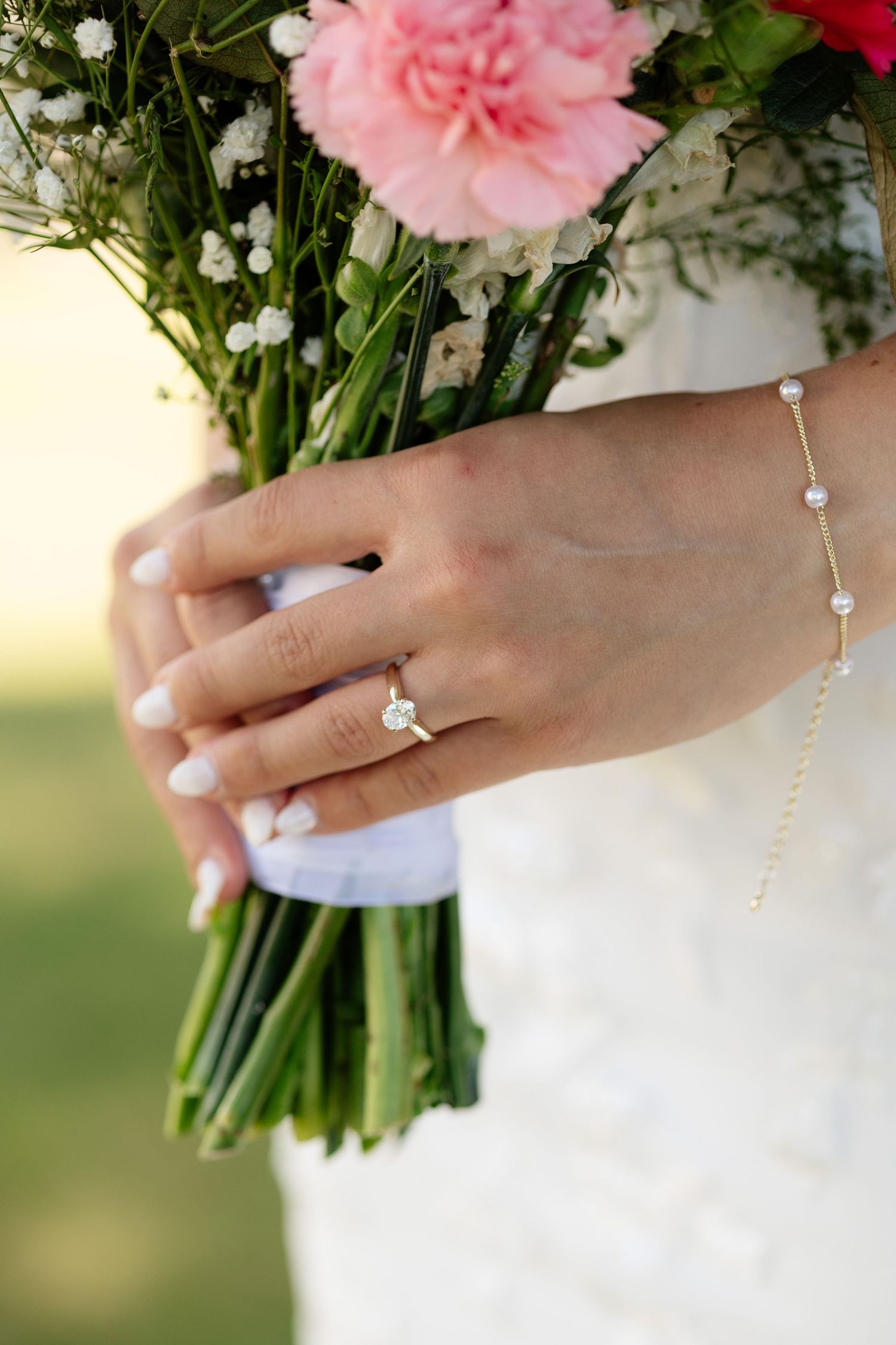 Bride holding pink floral bouquet with diamond solitaire engagement ring and delicate pearl bracelet on wedding day.