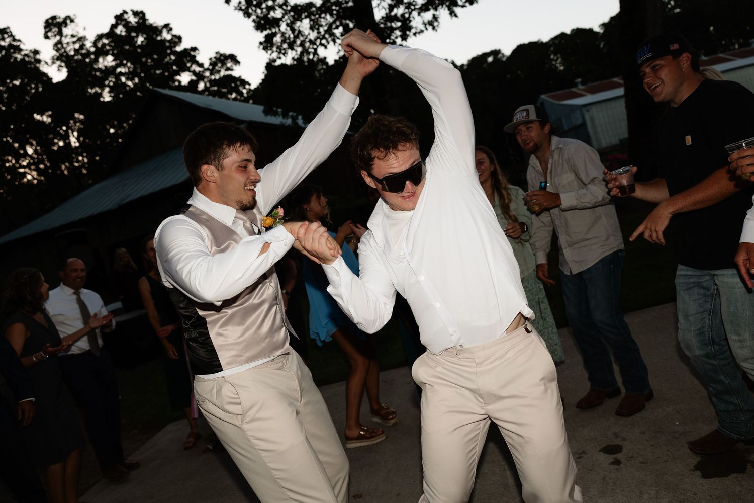Two men dancing energetically at outdoor wedding reception at dusk with guests in background.
