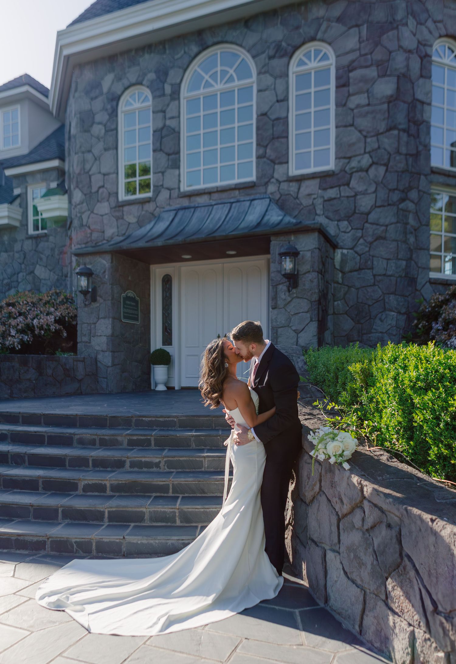 Two newlyweds embrace on stone steps outside an elegant stone mansion with arched windows on a sunny day.